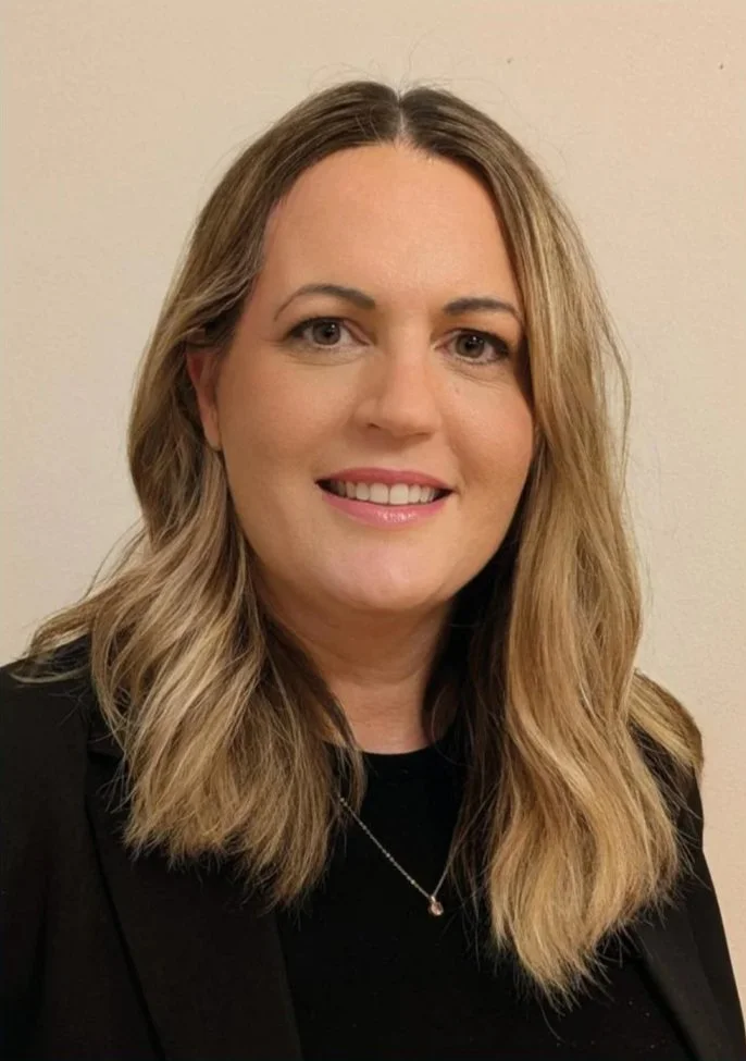 Ein Porträt einer Frau mit langen, gewellten, braunen Haaren, lächelnd vor einem hellen Hintergrund.