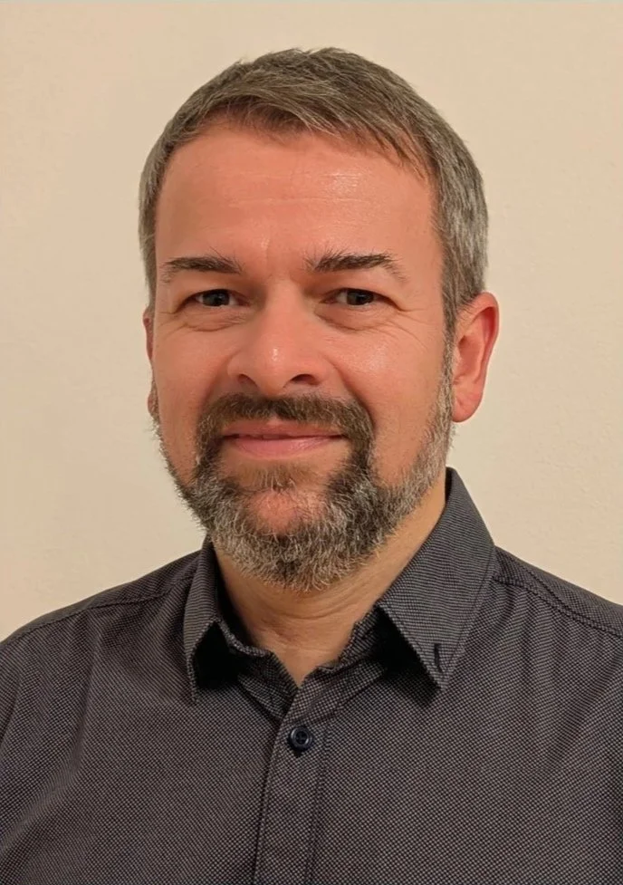 Ein Mann mit braunem Haar, braunem Bart und blauem Hemd, der vor einer weißen Wand steht.