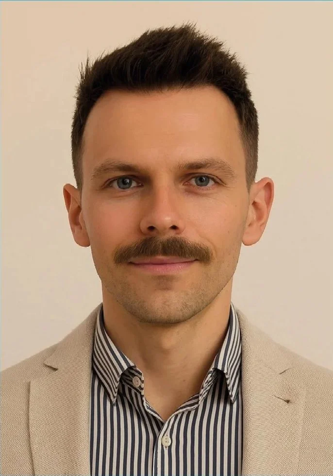 Ein Mann mit kurzer dunkler Frisur, Bart und Schnurrbart, trägt ein beige Sakko und ein gestreiftes Hemd, schaut direkt in die Kamera, vor einem neutralen Hintergrund.