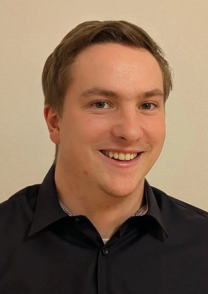 Ein Junge mit braunem Haar, der lächelt, trägt ein schwarzes Hemd vor einer hellen Wand.