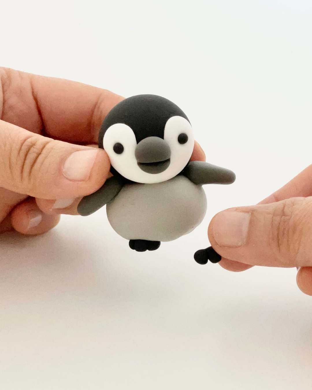 Hand holding a small penguin figurine made of clay or plastic, with a black head, white face, and gray body, against a plain white background.