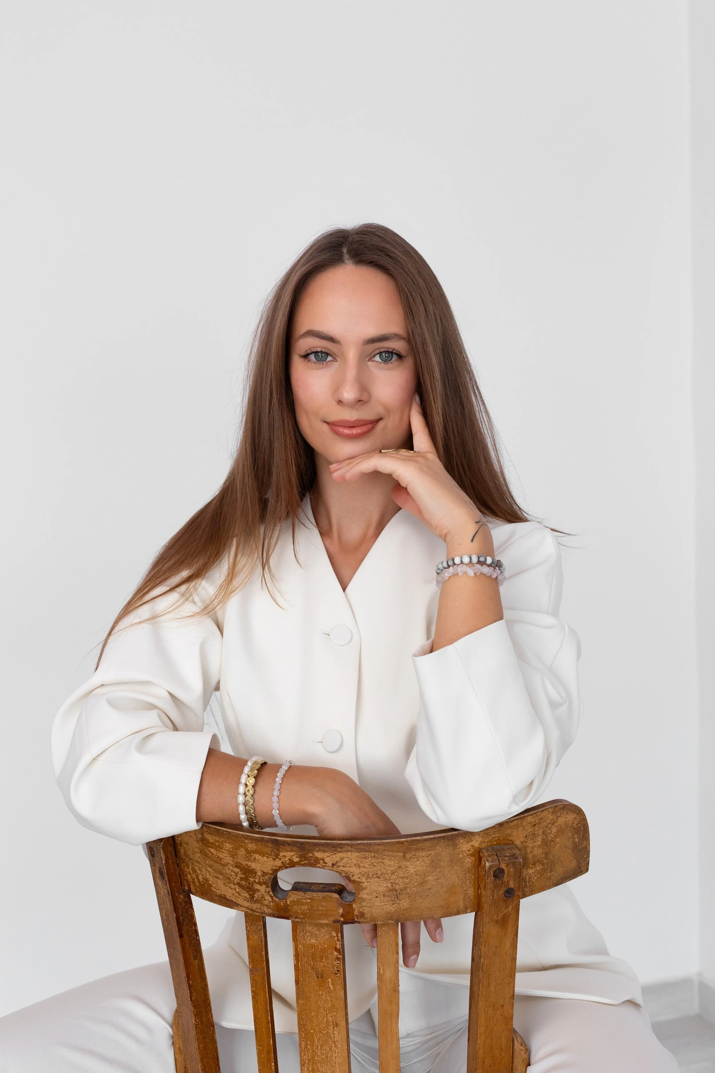 A young woman with long brown hair and blue eyes, sitting behind a wooden chair, wearing a white blazer and multiple bracelets, looking at the camera with a slight smile in a plain white room.