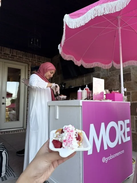 Person holding a plate of ice cream with strawberries in front of an ice cream stand with pink decorations and a large pink umbrella.