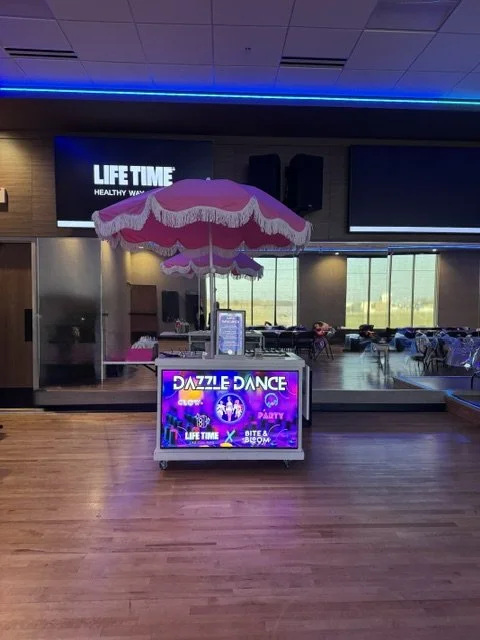 Indoor entertainment area with a pink and white canopy umbrella, a colorful Dazzle Dance booth, and chairs in the background.