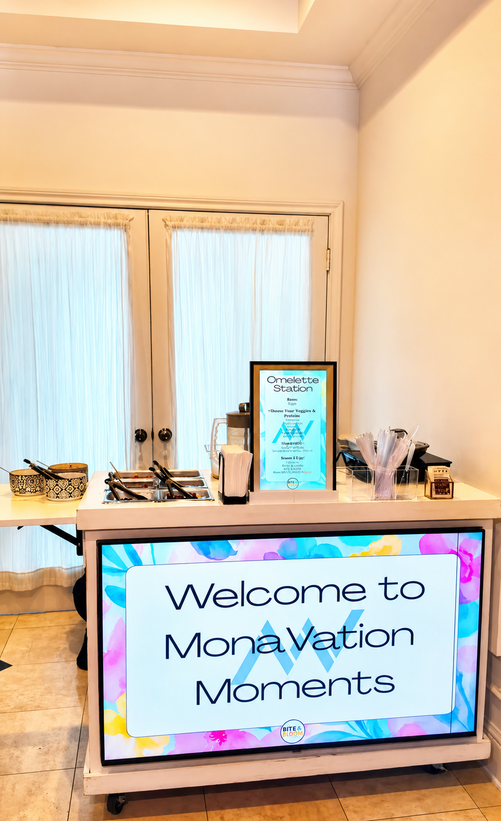 A welcome station at MonaVation Moments with a digital sign, buffet area, and utensils, set up in a bright room with curtains.