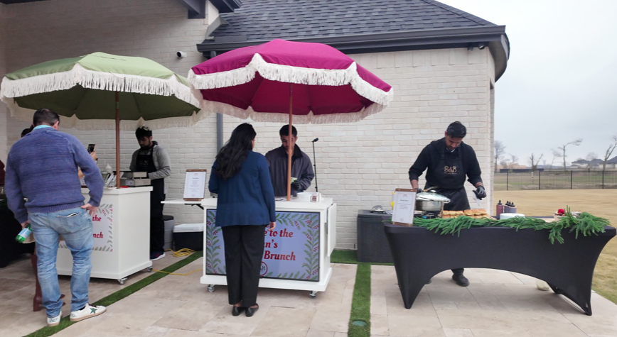 People at an outdoor brunch buffet under two large umbrellas, with servers preparing food and guests waiting in line.