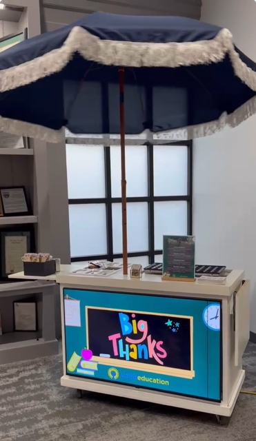 A classroom-style information booth with a large blue umbrella on top, featuring a sign that reads 'Big Thanks' and 'education,' displayed in a room with shelves and windows.