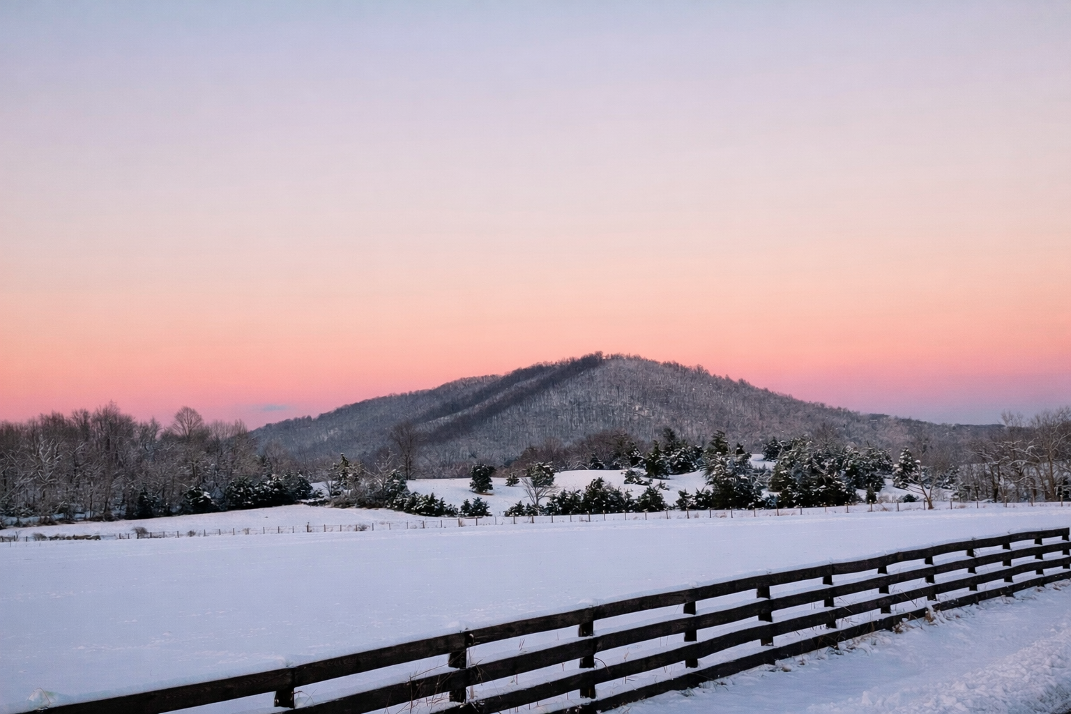 Snow-covered field with a wooden fence in the foreground, leafless trees and snow-covered bushes in the middle, and a mountain in the background under a pastel pink and blue sky at dusk.