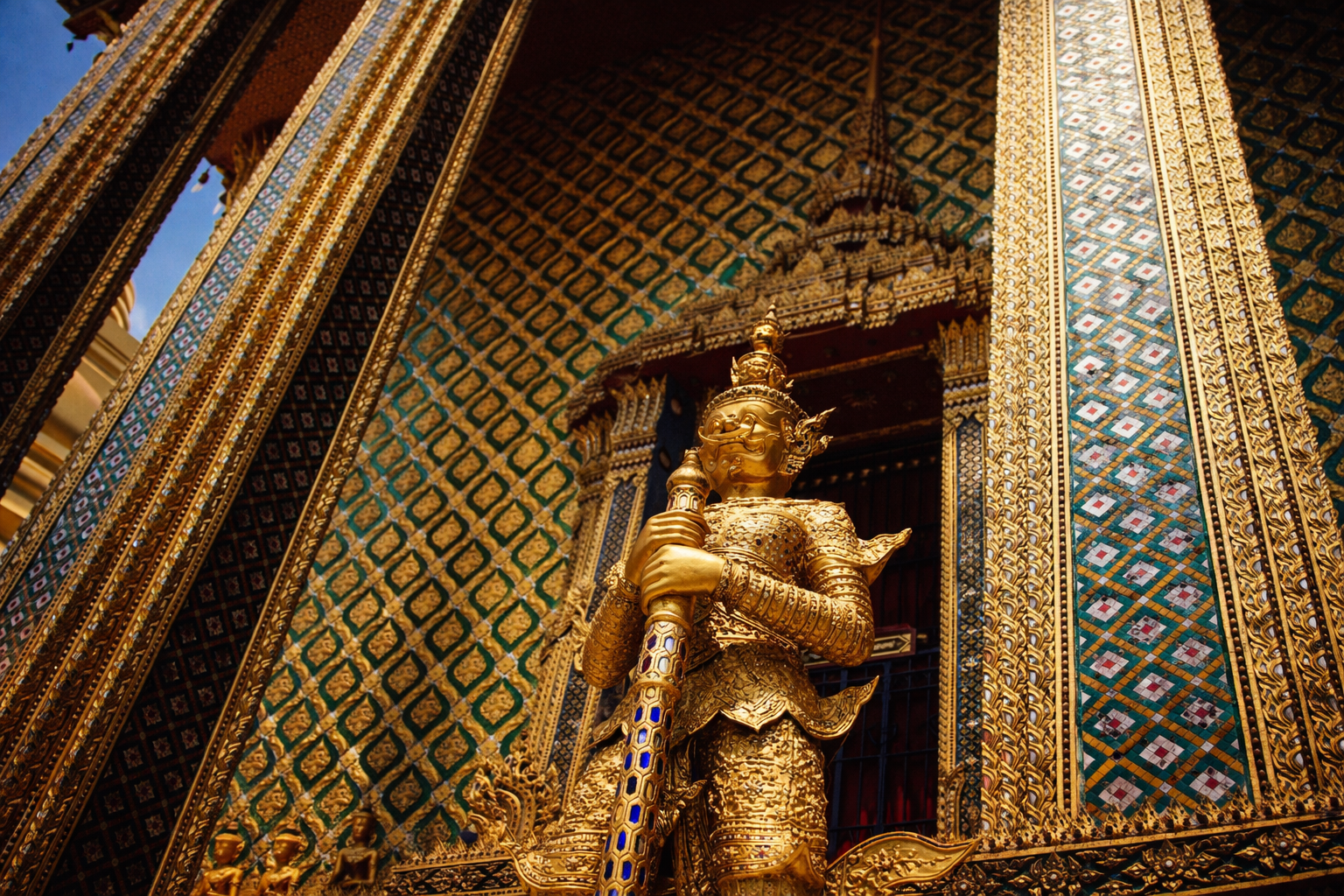 Close-up of a golden sculpture of a mythical guardian figure holding a sword, set against an ornate, patterned temple wall with gold, blue, and red accents.