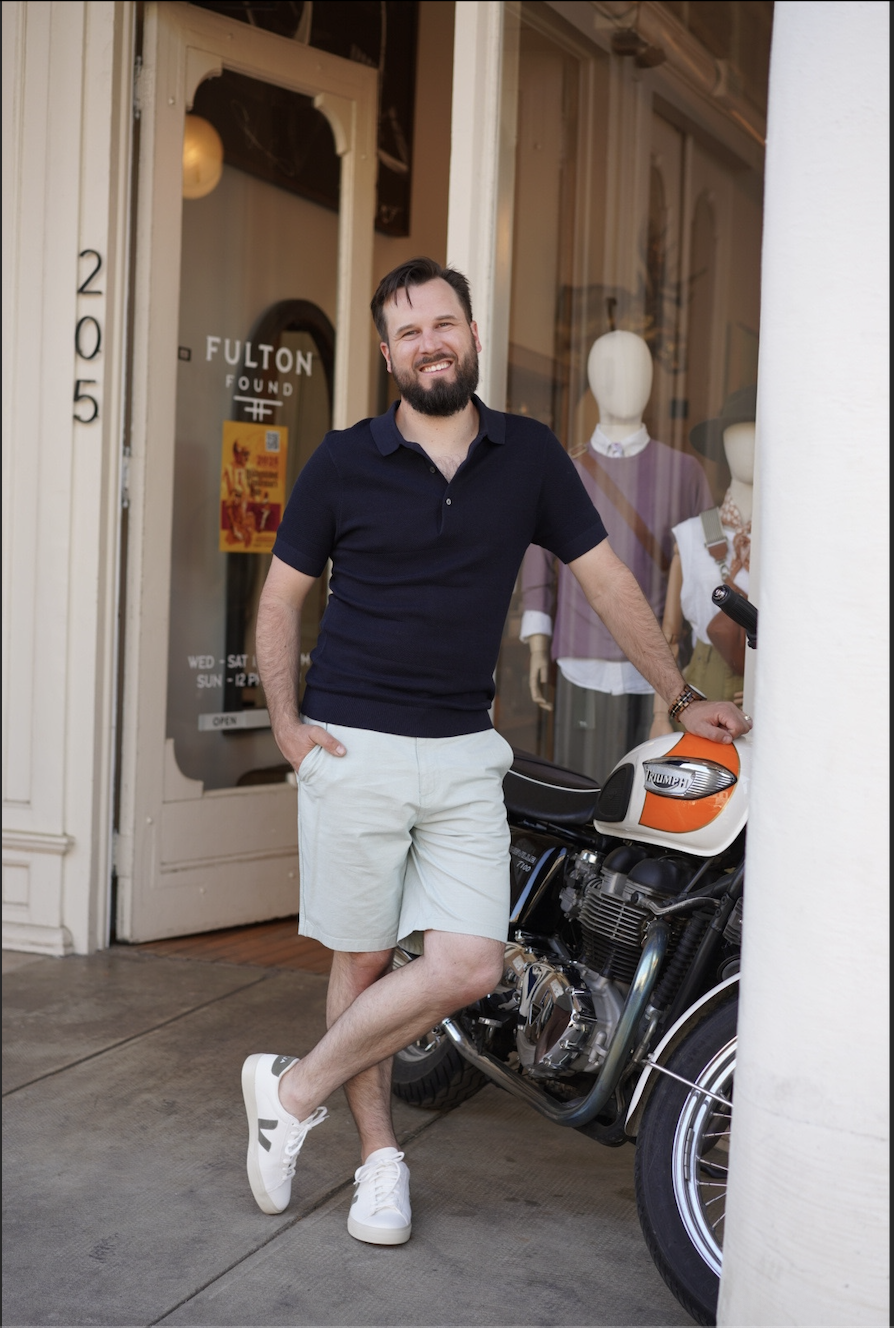 A bearded man in a navy polo shirt and white shorts standing outside a storefront, leaning on a vintage motorcycle, smiling at the camera.