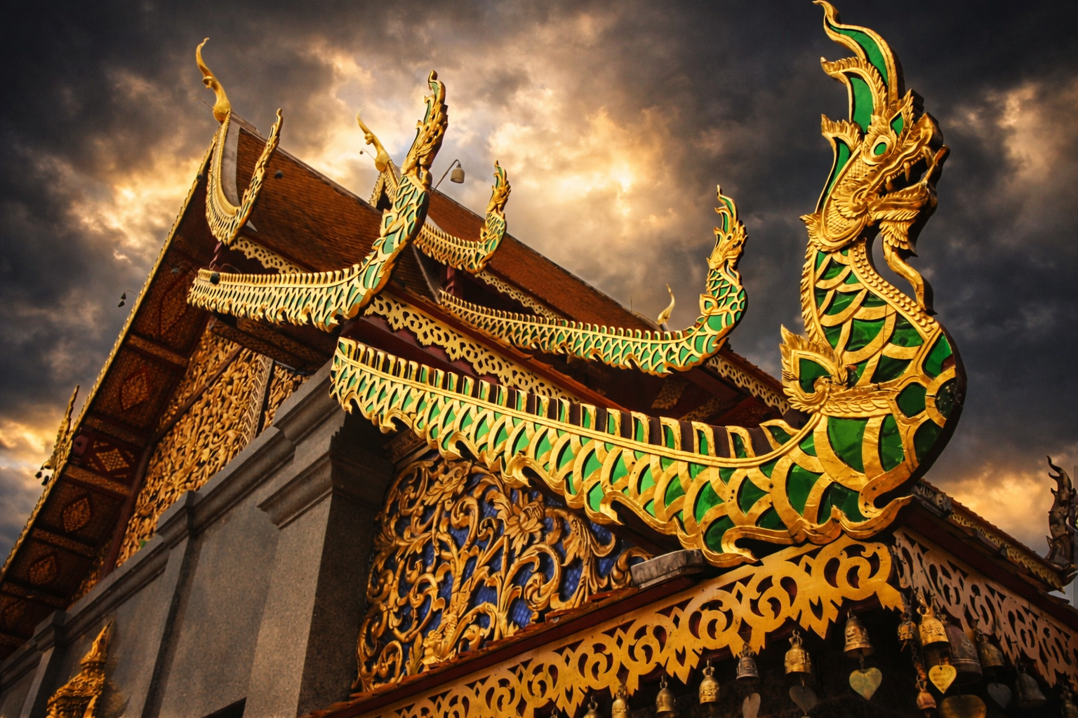 Ornate Thai temple roof with golden and green dragon-like decorative elements against a dramatic cloudy sky.