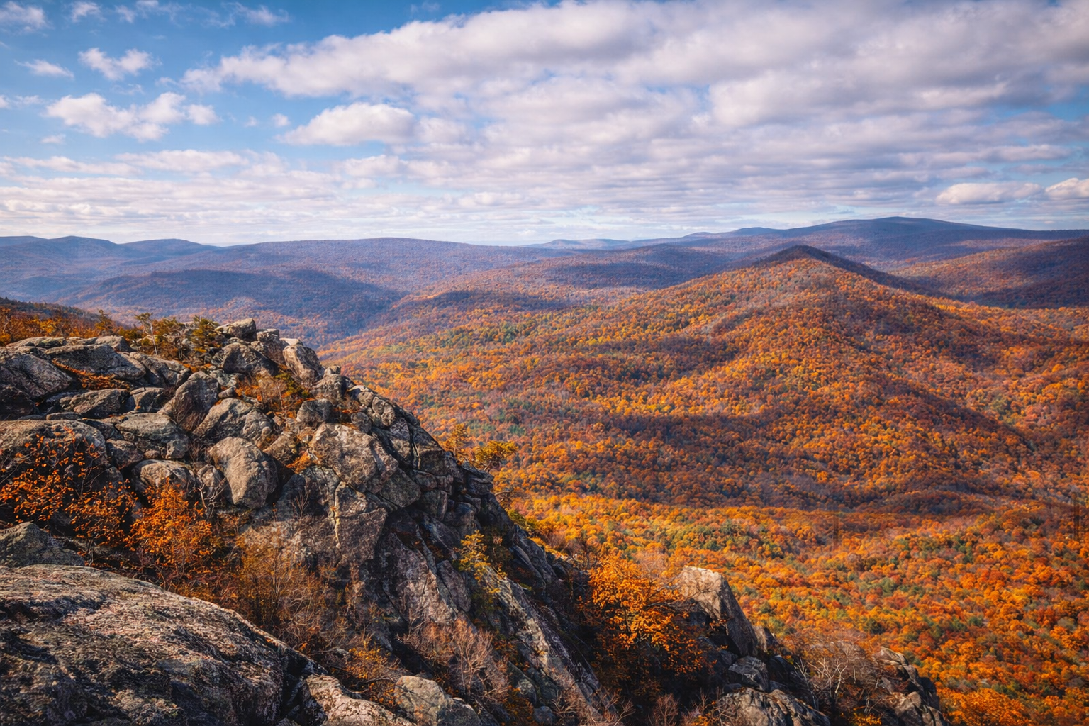 Mountain landscape with colorful autumn foliage, rocky foreground, and a partly cloudy sky.