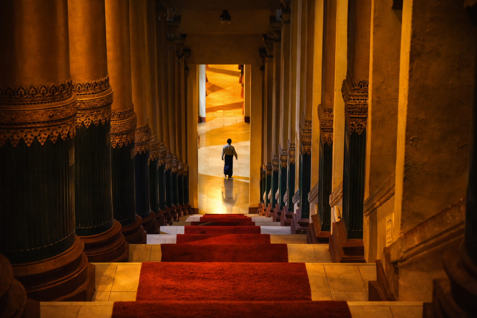 View of a staircase with red carpet runners, ornate columns, and warm yellow lighting, leading down towards a person walking away in the distance.