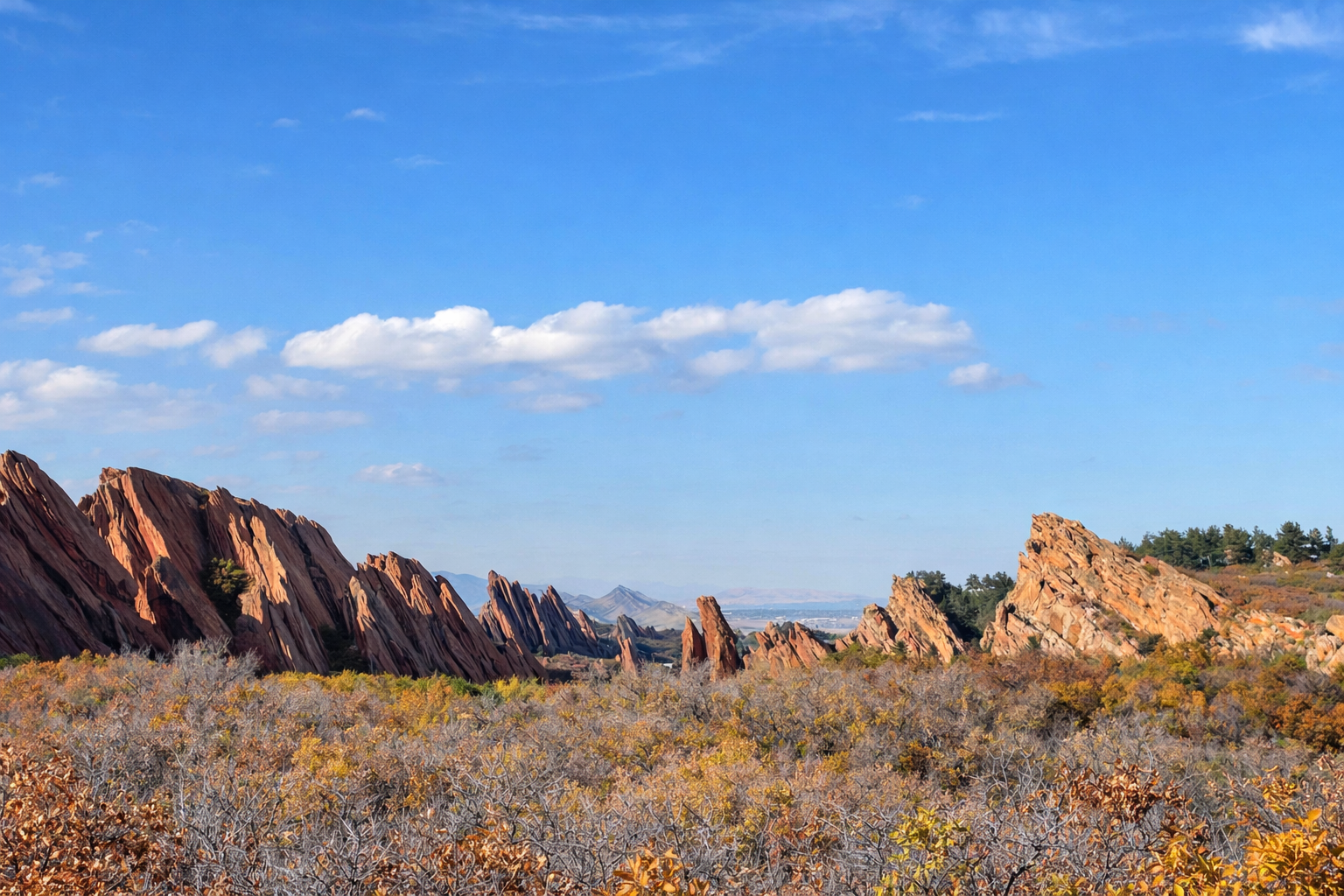 Scenic desert landscape with jagged reddish-brown rock formations under a blue sky with scattered clouds, and autumn-colored bushes in the foreground.