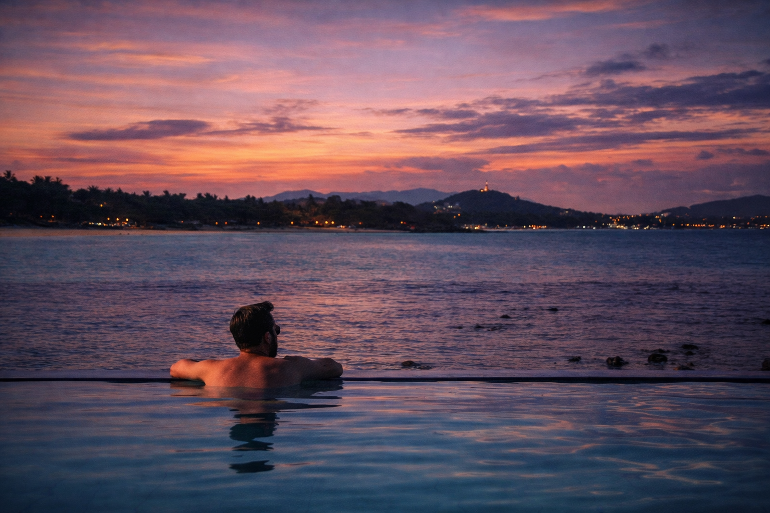 A man with dark hair and a beard is relaxing in an infinity pool during sunset, overlooking the ocean with mountains and a cityscape in the distance, with a colorful sky of pink, purple, and orange hues.