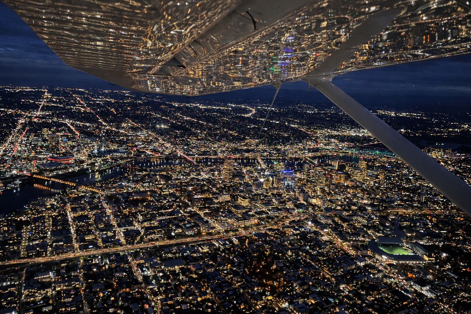 Aerial view of a city at night taken from an airplane, showing illuminated streets, buildings, and a river, with the airplane wing and strut visible in the foreground.