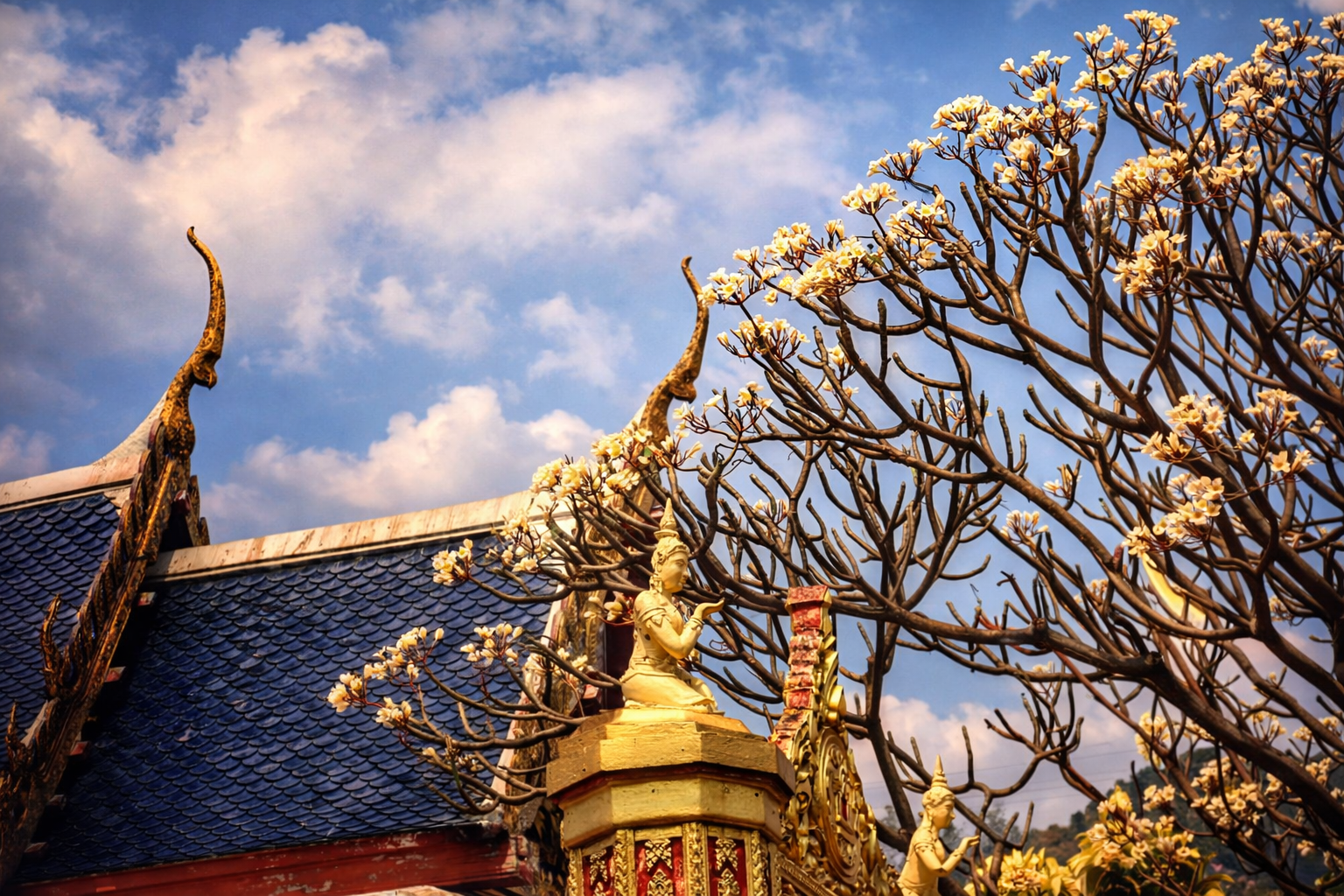Temple roof with ornate golden sculptures and a tree with white blossoms against a blue sky.