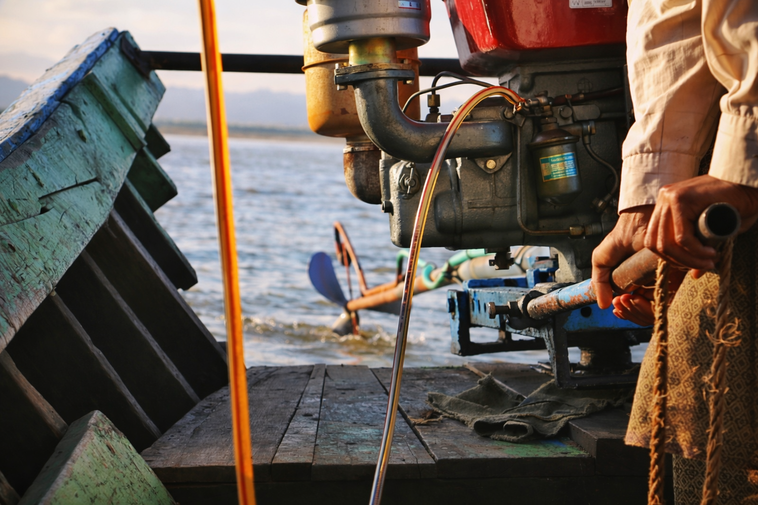 Close-up of a person's hand holding a tool near a boat's engine, with a body of water and another boat in the background.