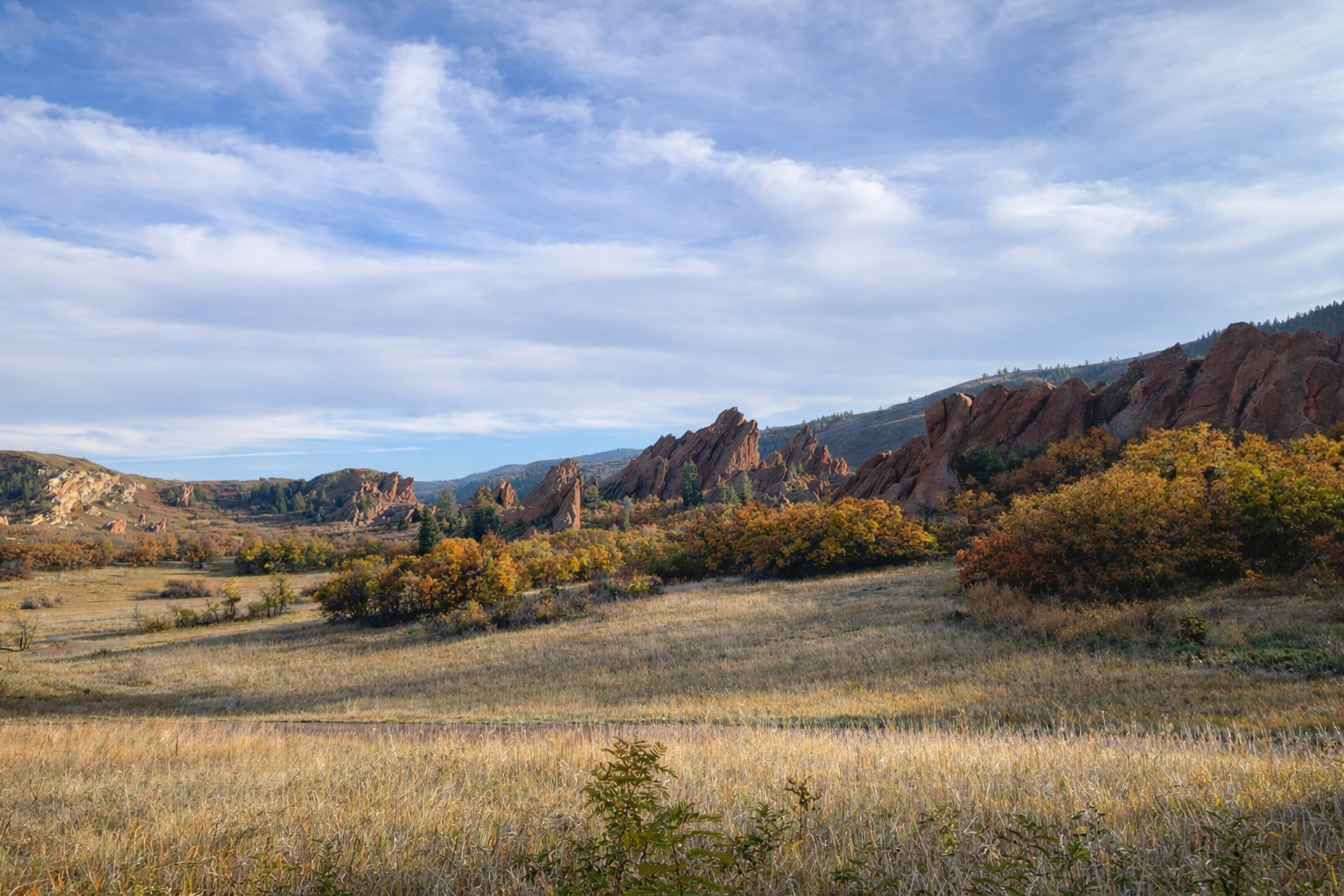 Scenic landscape of flat grassy field with yellow and orange trees, rocky mountain formations in the background, and a partly cloudy sky.