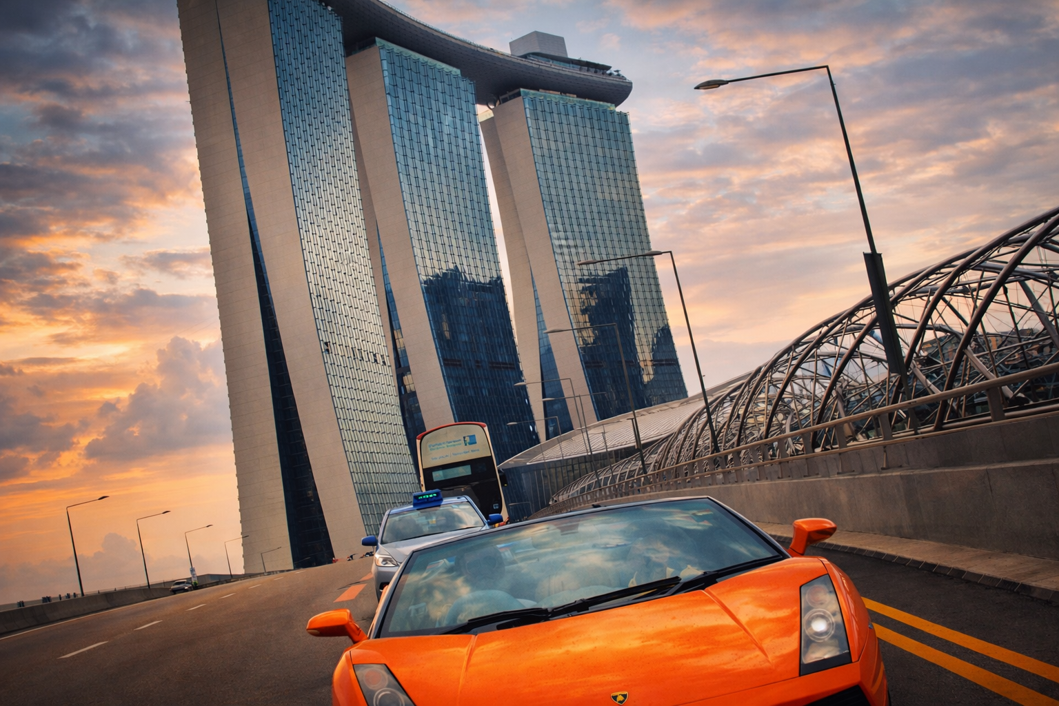 A street scene in front of a modern high-rise building with reflective glass windows during sunset. An orange sports car and police cars are on the road, with street lamps lining the street.