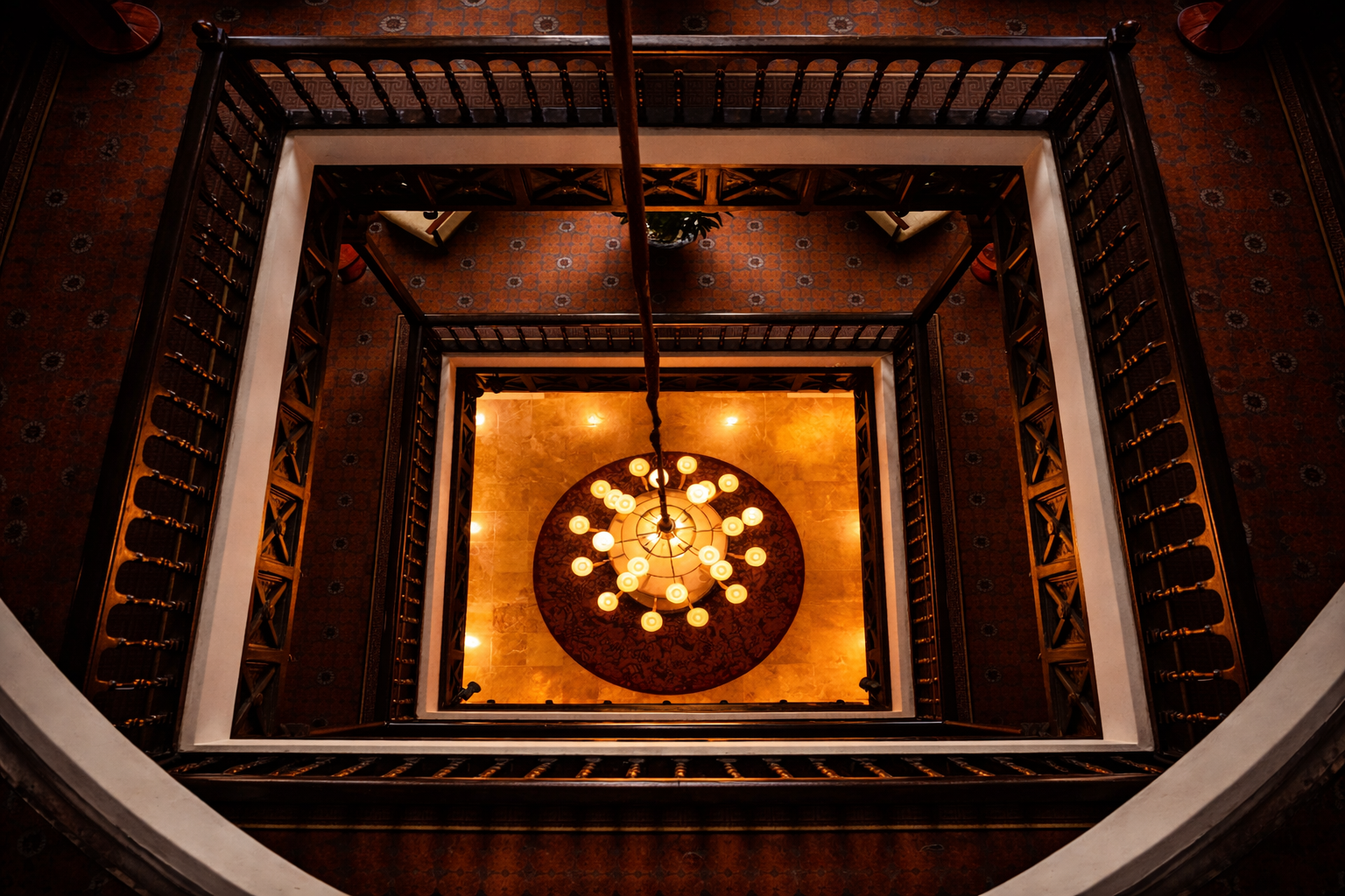 Looking down at a multi-tiered chandelier hanging over a spiral staircase with dark wood railings in a hotel lobby.