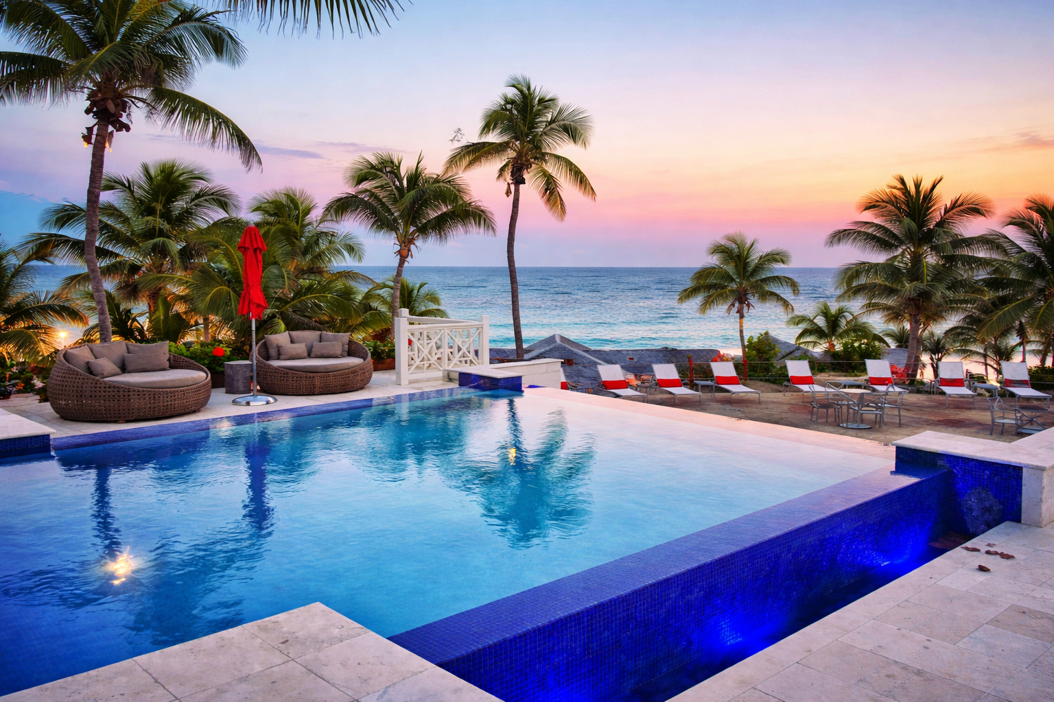 A tropical beachside pool area at sunset with palm trees, lounge chairs with red and white cushions, wicker lounge chairs with cushions, and a view of the ocean and colorful sky.