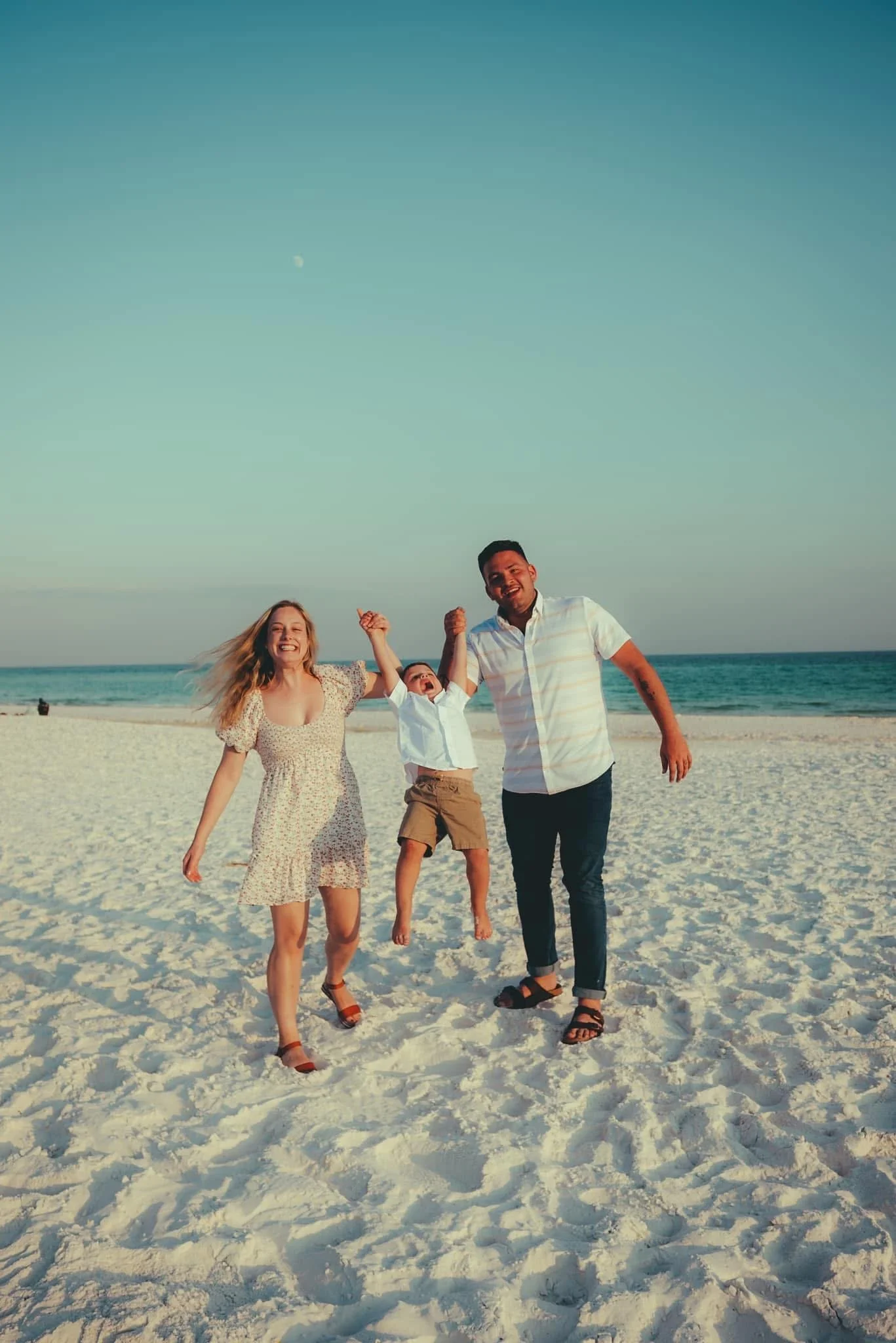 A family of three enjoying a day at the beach, with the mother, father, and child holding hands and jumping in the air on white sand with ocean waves in the background.