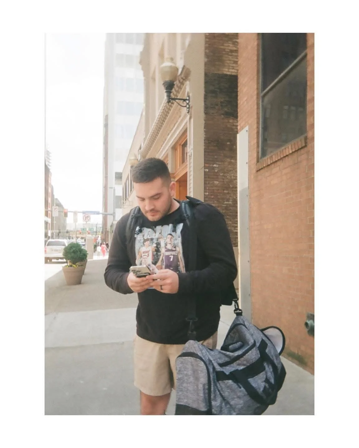 A young man with short dark hair, wearing a graphic t-shirt and beige shorts, looking at his phone while standing outside on a city sidewalk. He has a backpack on his back and a gray duffle bag on the ground beside him. The background features brick and stone buildings, a potted plant, and street activity.