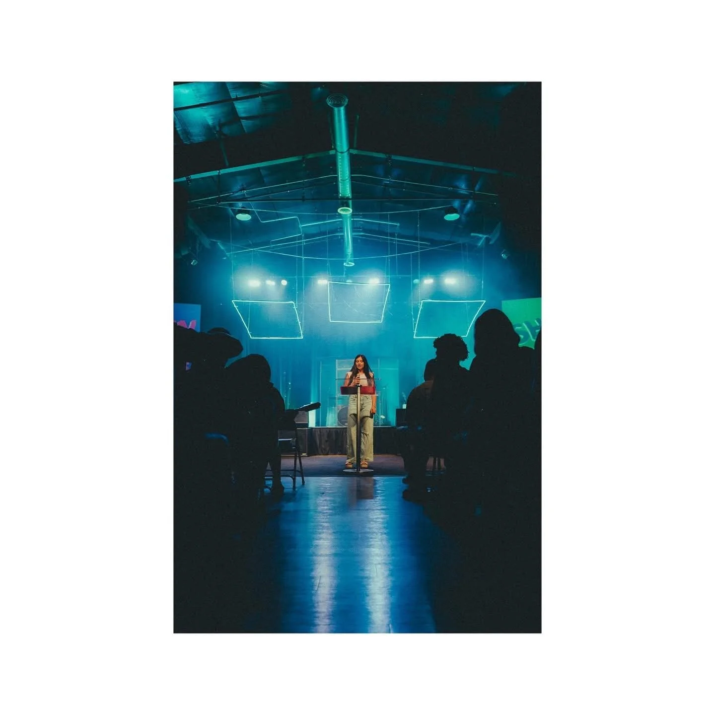 A woman performs on stage in a dark auditorium with blue lighting and geometric light fixtures overhead, while audience members sit in silhouette.