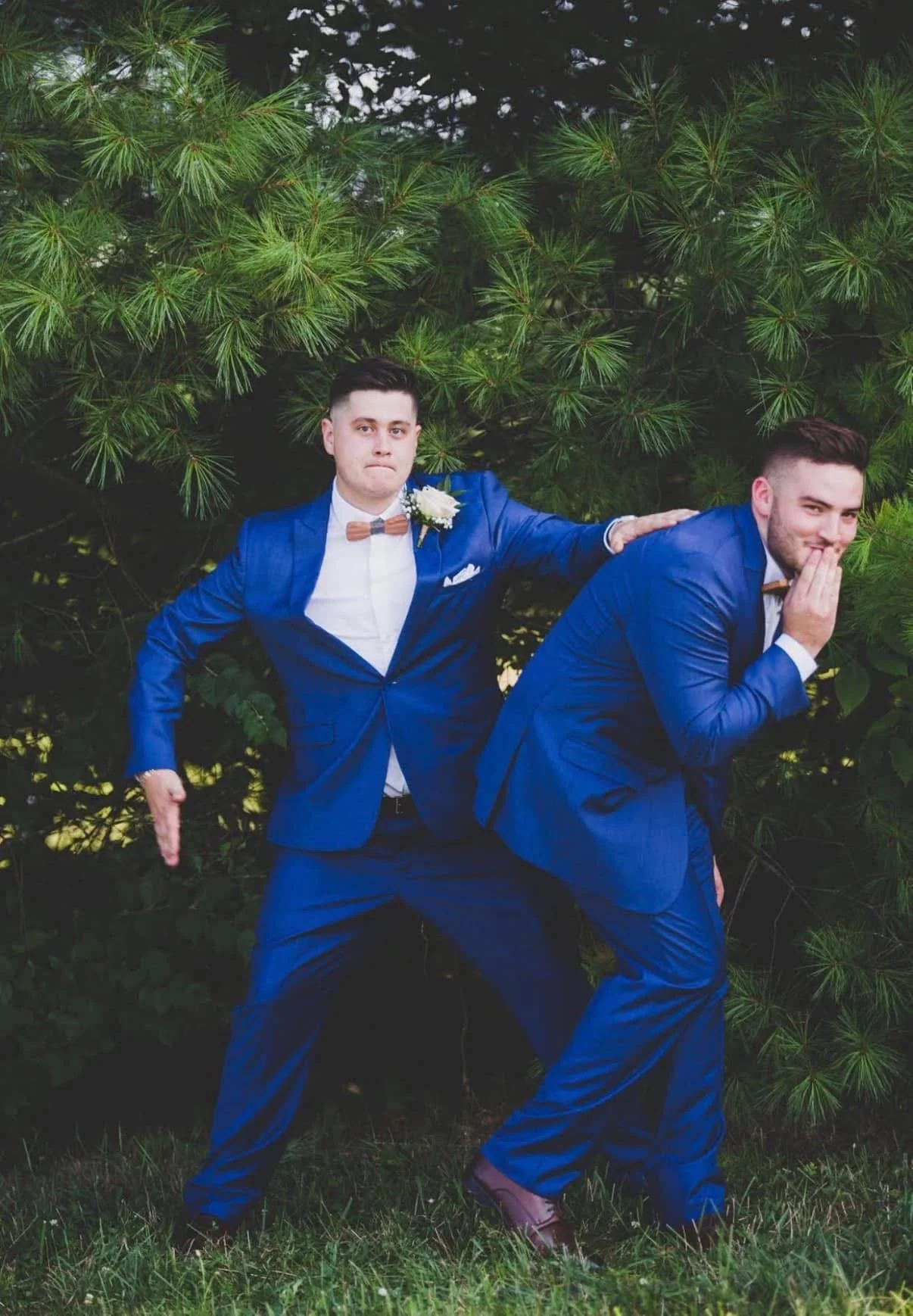Two men in blue suits posing outdoors in front of green trees, one with a surprised expression and the other with a playful gesture.