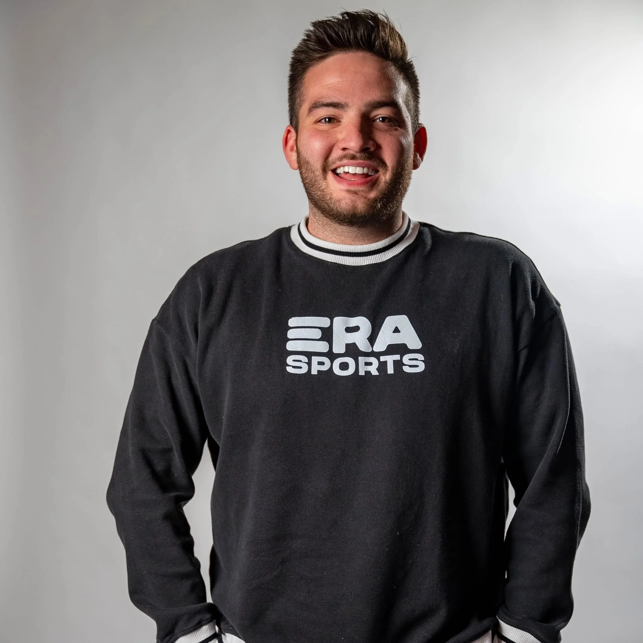 A young man with short, styled brown hair and facial hair, smiling, wearing a black sweatshirt with white trim and a logo that says "ERA SPORTS" on a gray background.