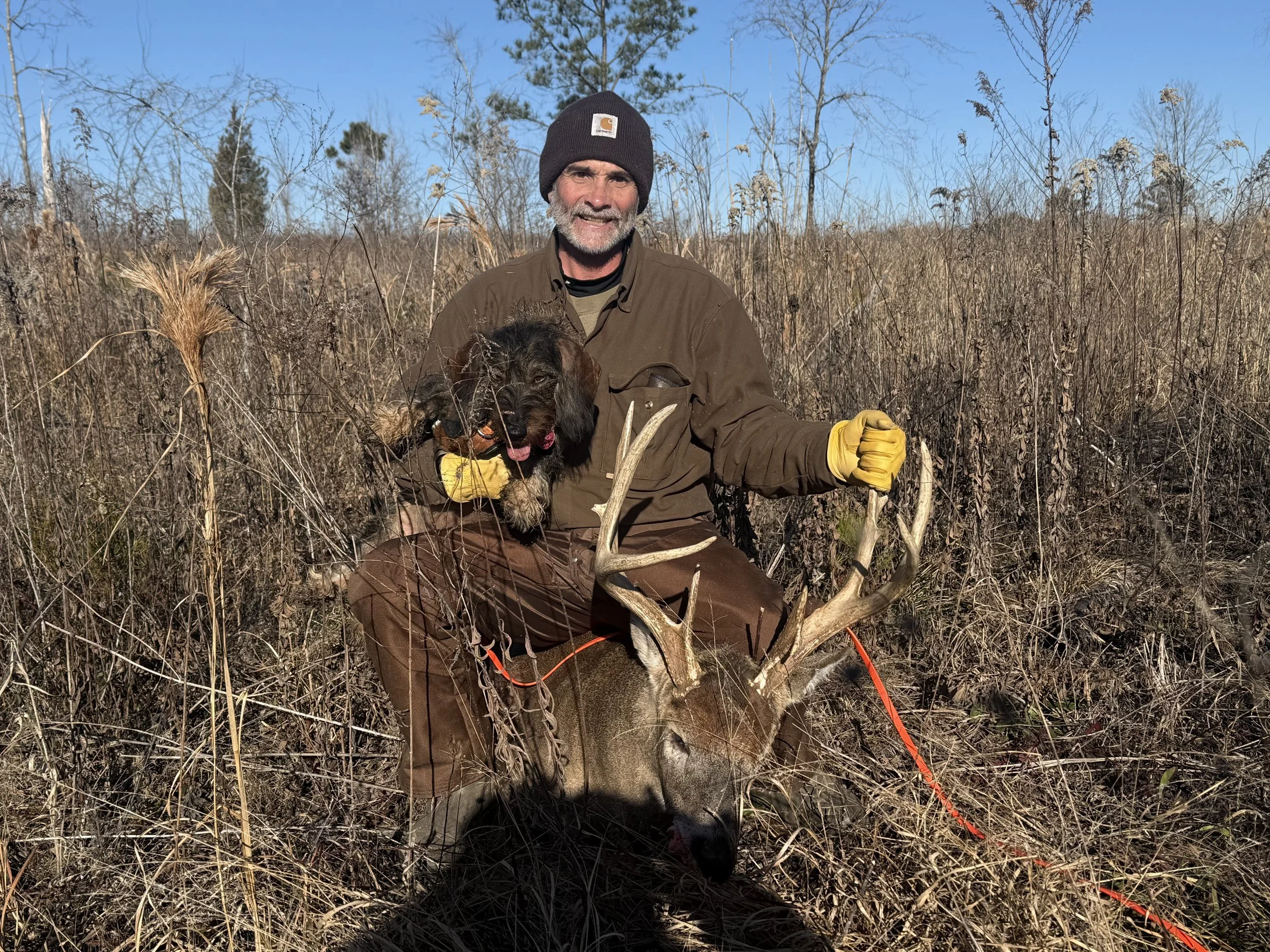 A man in outdoor clothing and yellow gloves holding a dog sitting on the back of a large deer with antlers, in a field of dry grass and shrubs on a clear day.