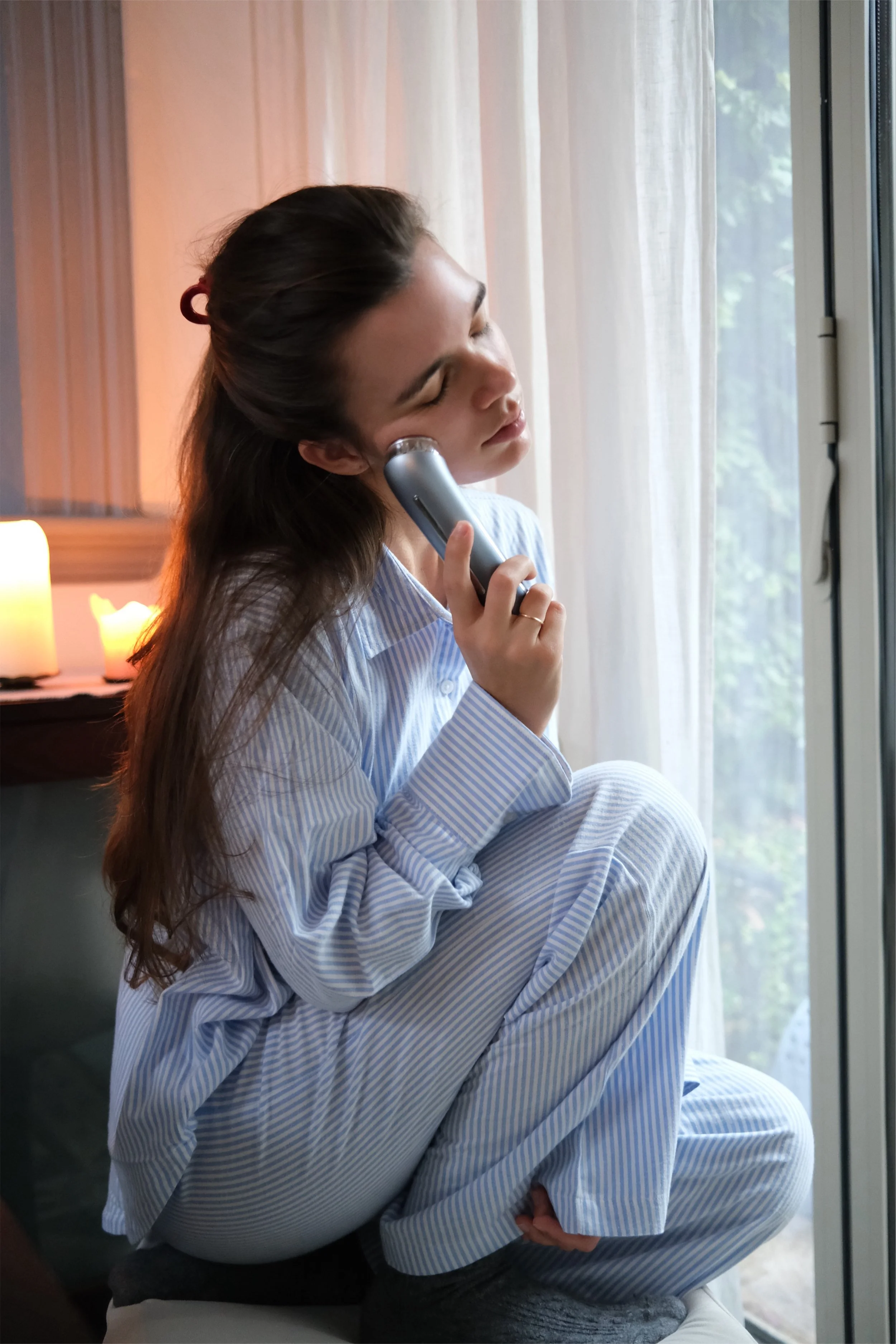 A young woman with long brown hair in pajamas, sitting cross-legged on a stool, talking on a cordless phone near a window with sheer curtains, with cozy candles in the background.