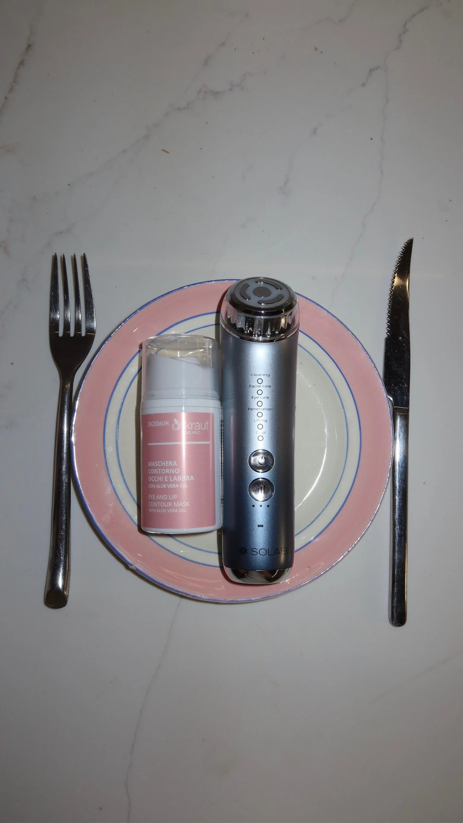 A small pink and white plate with silver fork and knife on white surface. On the plate are a silver skincare device and a pink bottle of eye and lip contour mask.