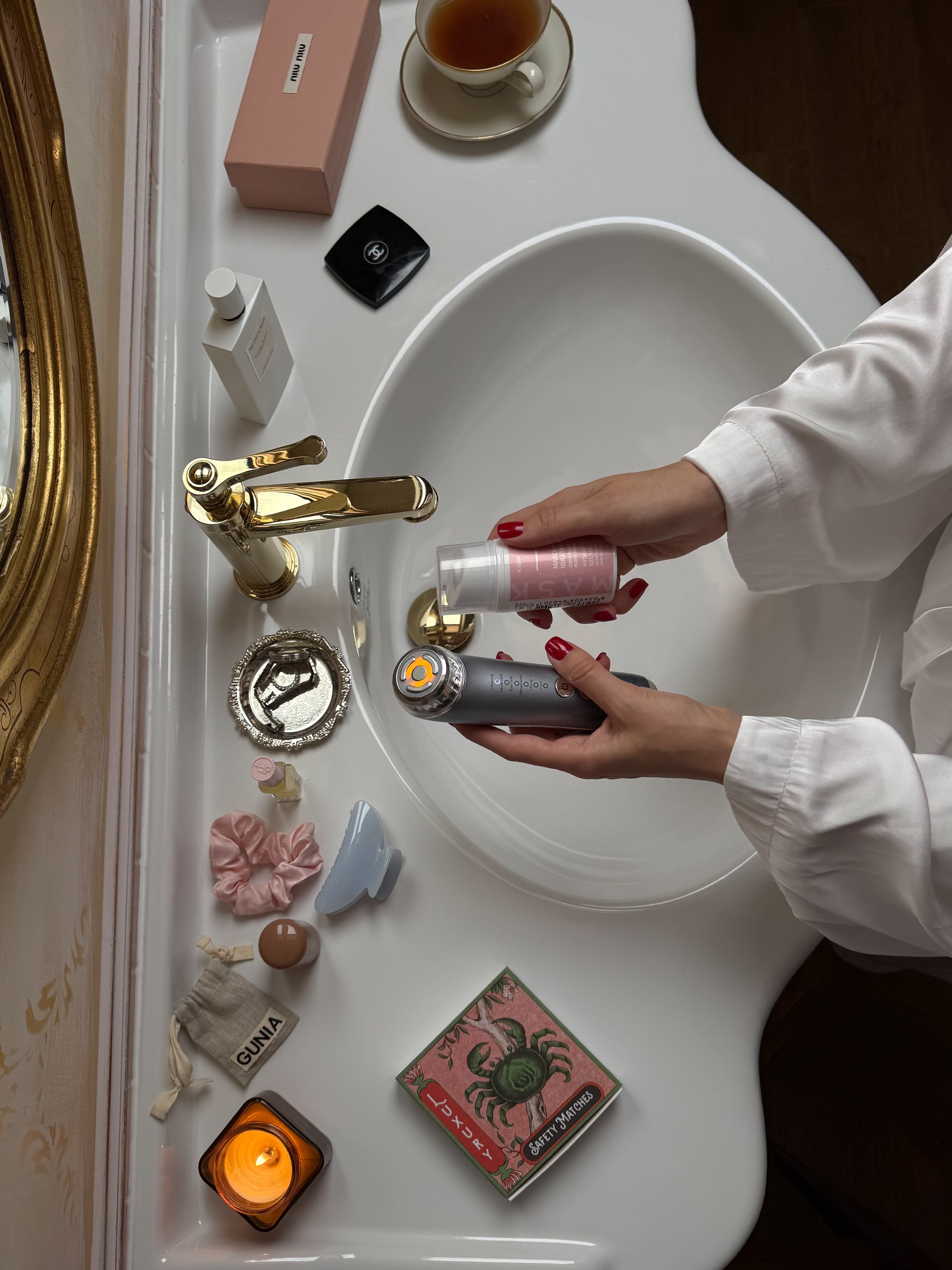 A white bathroom sink countertop with a gold faucet, various toiletries, a pink box, a cup of tea, and a person's hands holding a skincare product and a beauty device.