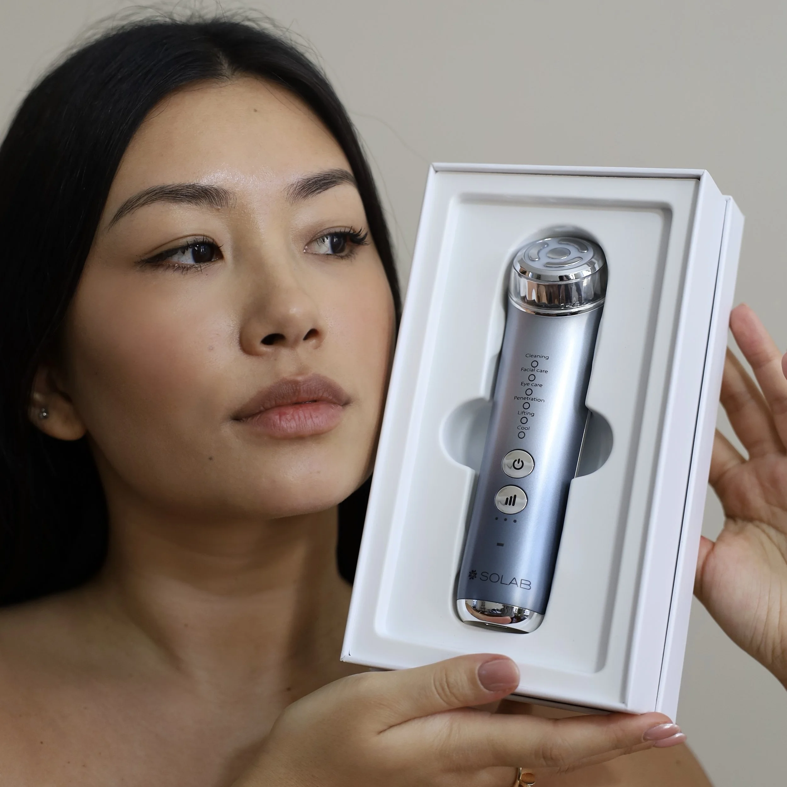 A woman with black hair holding up a box with a silver skincare device branded 'SOLAB' inside.