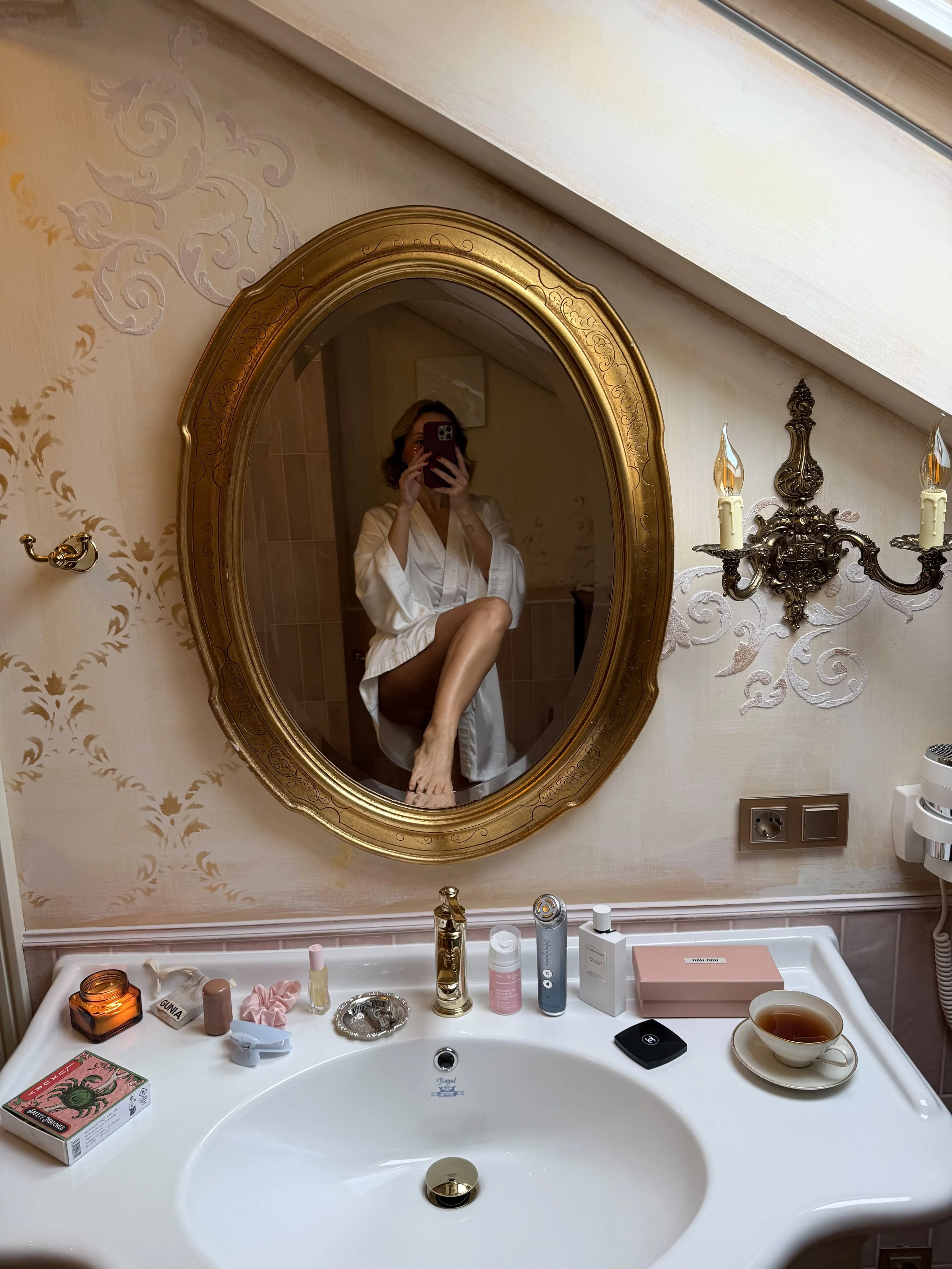 Woman in a white robe sitting on a bathroom sink with a gold-framed oval mirror behind her. She is taking a photo with her phone, with her legs crossed. The bathroom has a cream-colored wall with decorative patterns, a sconce with two candles, and va