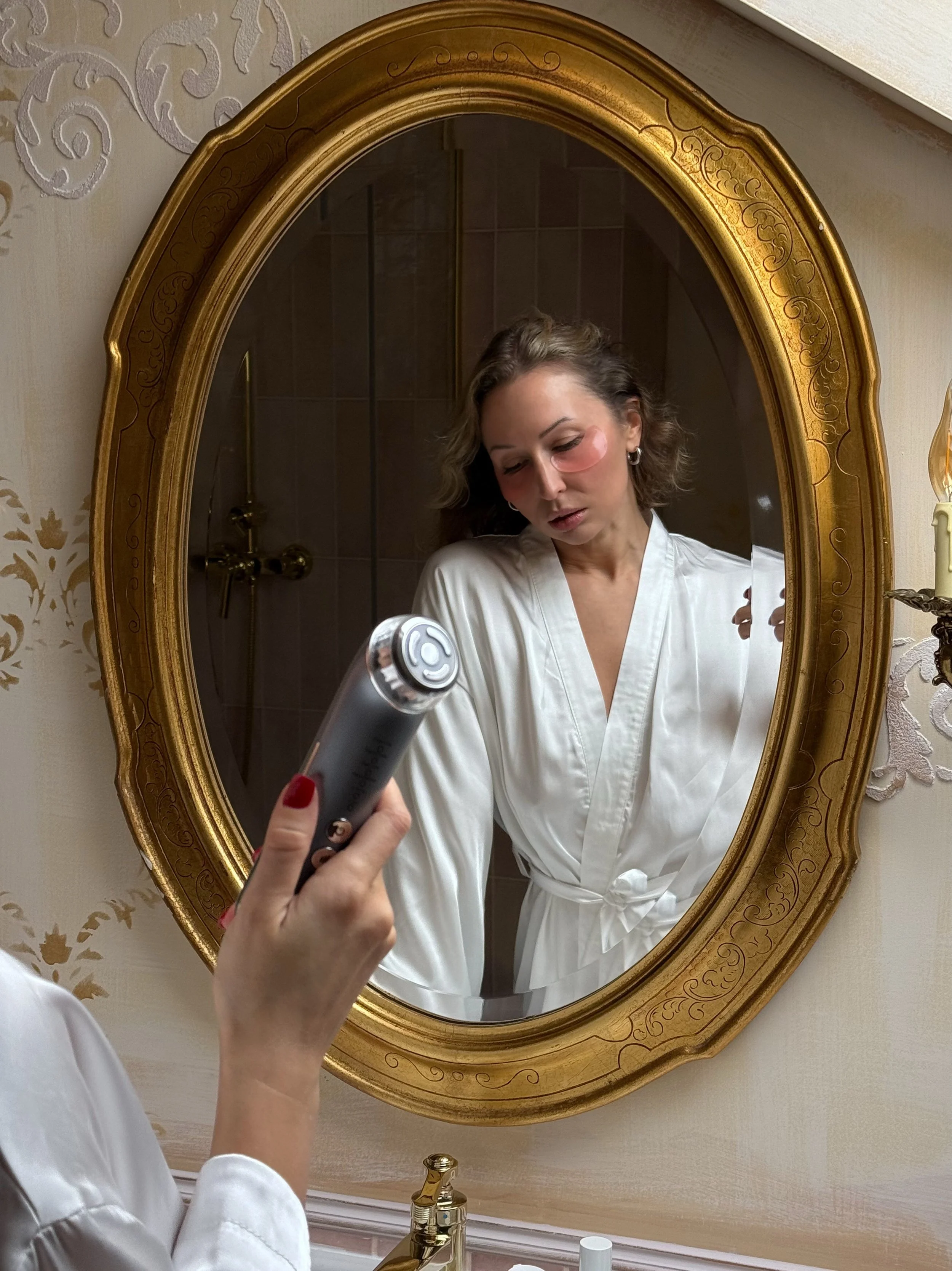 A woman in a white satin robe looking at herself in an ornate gold-framed mirror while someone holds a skin care device.