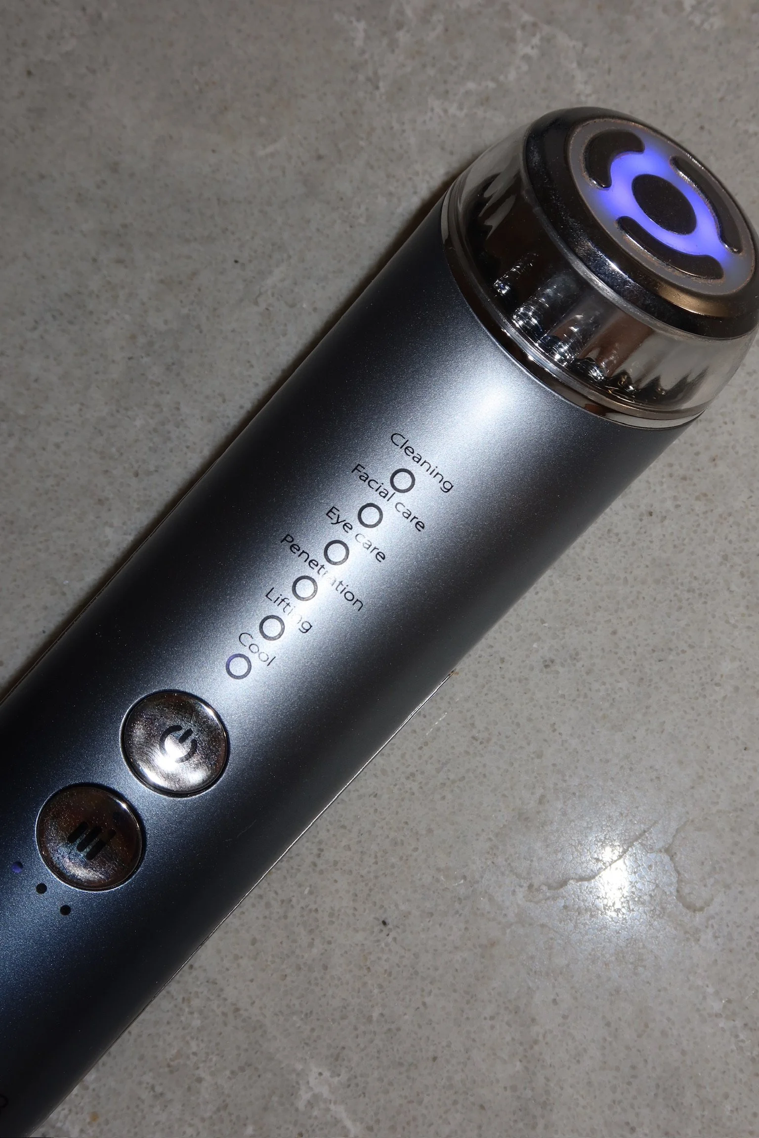 A silver facial cleansing device with a purple-lit attachment on one end, featuring three buttons on the side.