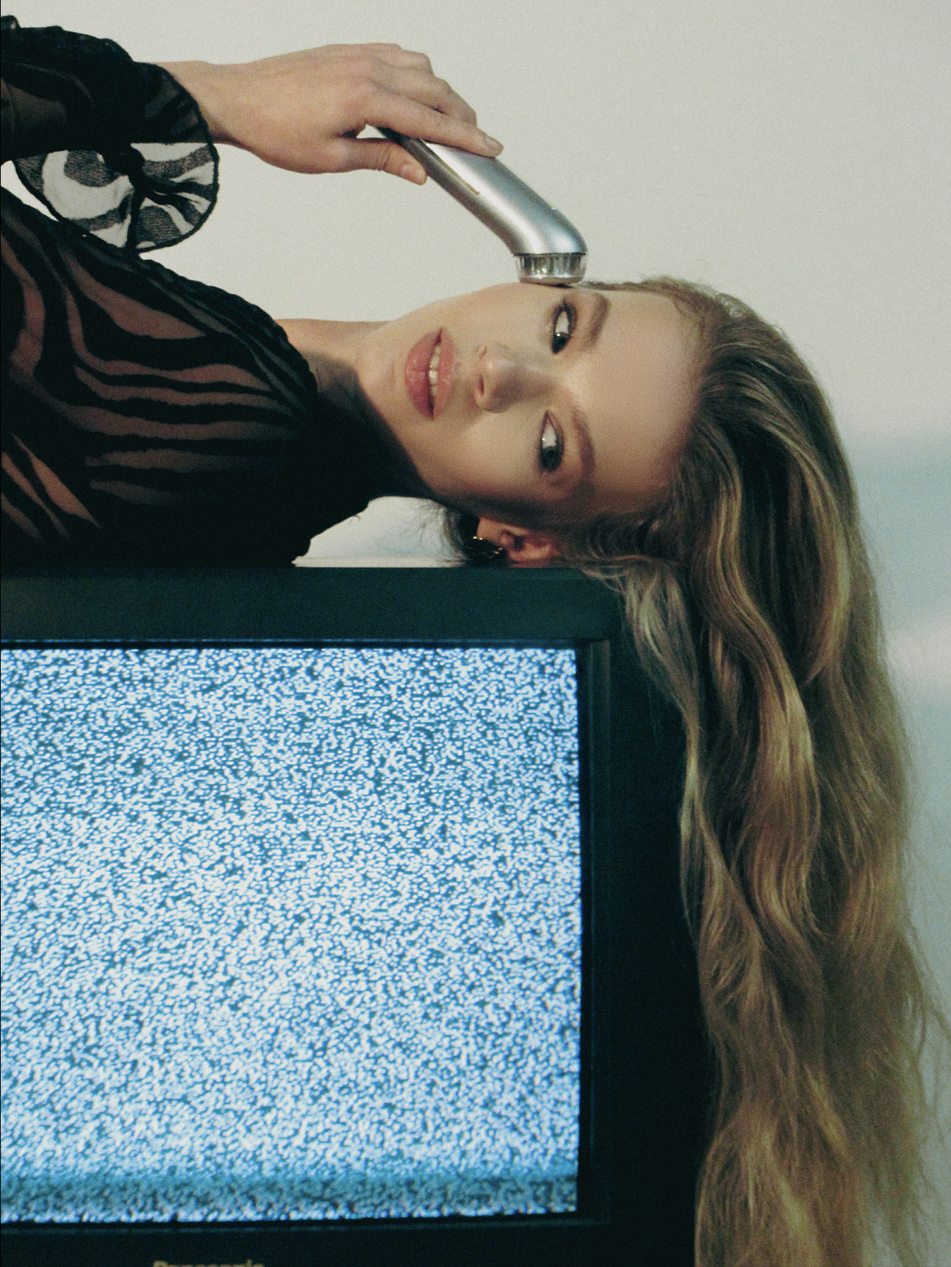 A woman with long wavy hair and a black, sheer, striped top is lying down with her head resting on a surface, holding a remote control to her head, while a television screen displaying static is in the foreground.