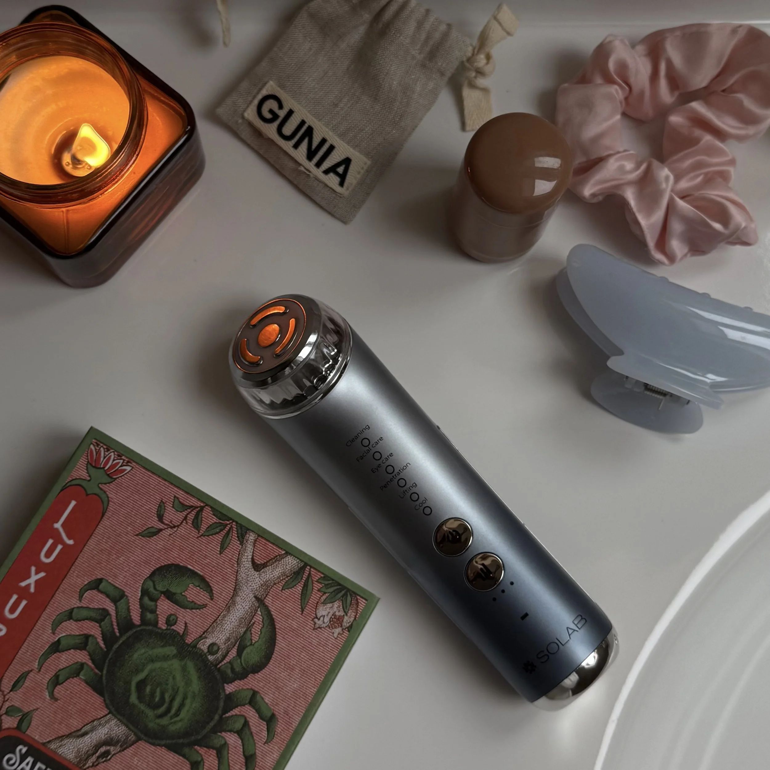 A collection of skincare and wellness items on a white surface, including a lit orange candle in a glass jar, a beige pouch labeled "GUNIA," a brown bottle, a pink scrunchie, a white facial steamer, a gray facial device, a box with an illustration of