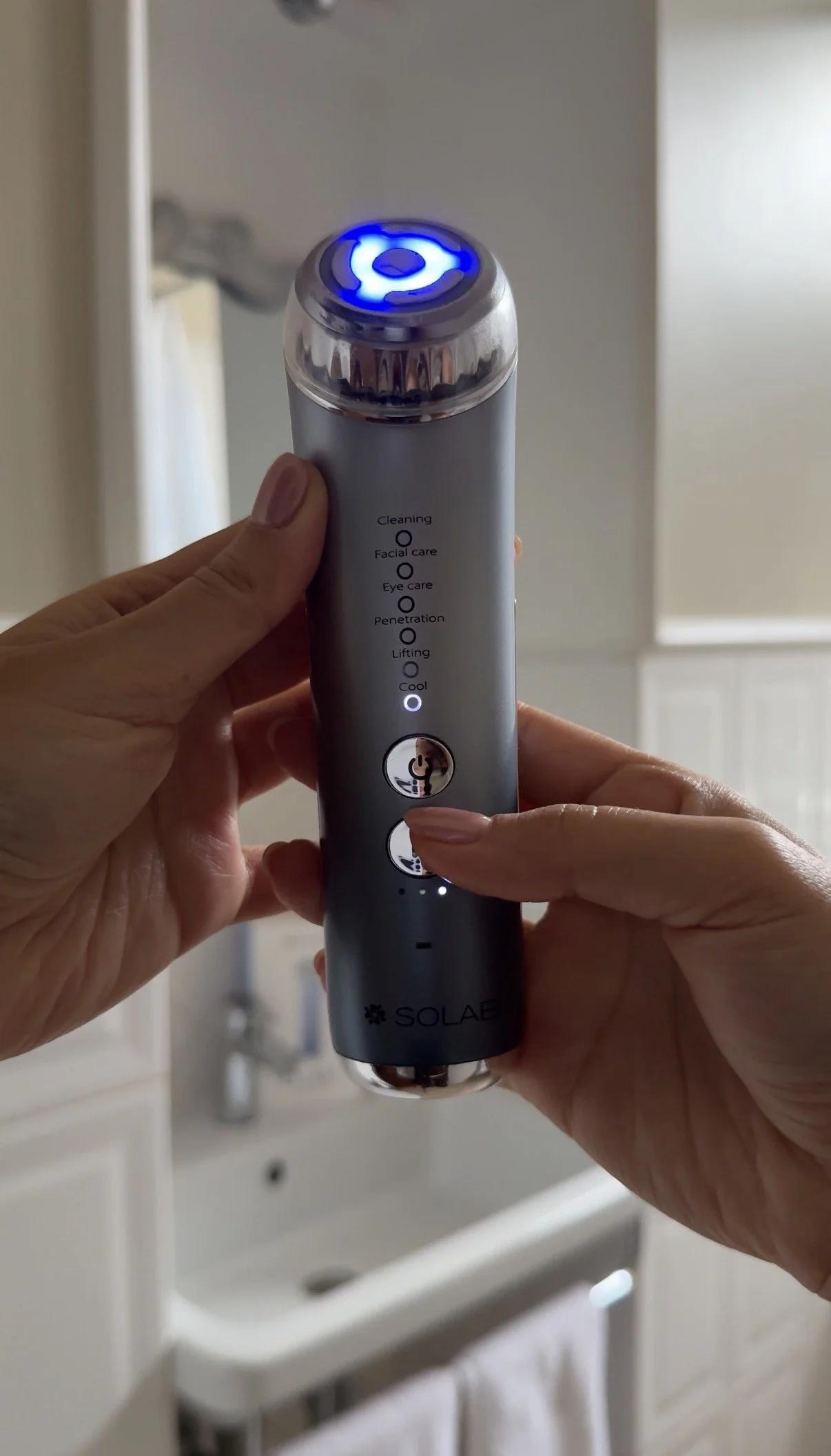 A person is holding a facial cleansing device with a blue LED light on top. The device has options listed vertically: Cleaning, Facial care, Eye care, Penetration, Lifting, Cool. The person is pressing a button on the device, which is branded 'Solae,