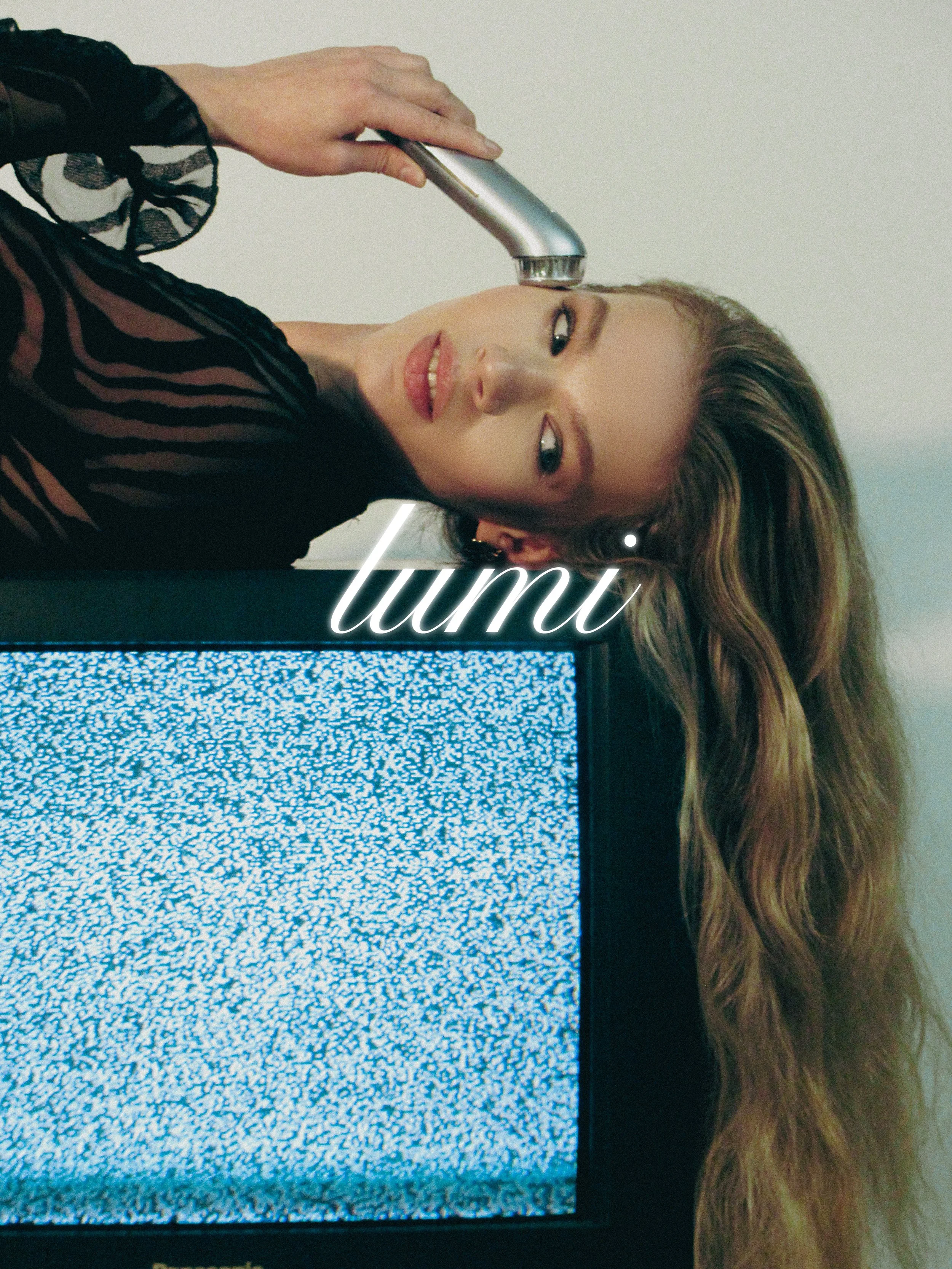 A woman with long wavy hair and a black sheer top is lying sideways next to an old television displaying static. She is holding a silver vintage microphone close to her mouth, and the word 'tumi' is written across the image in elegant white cursive.