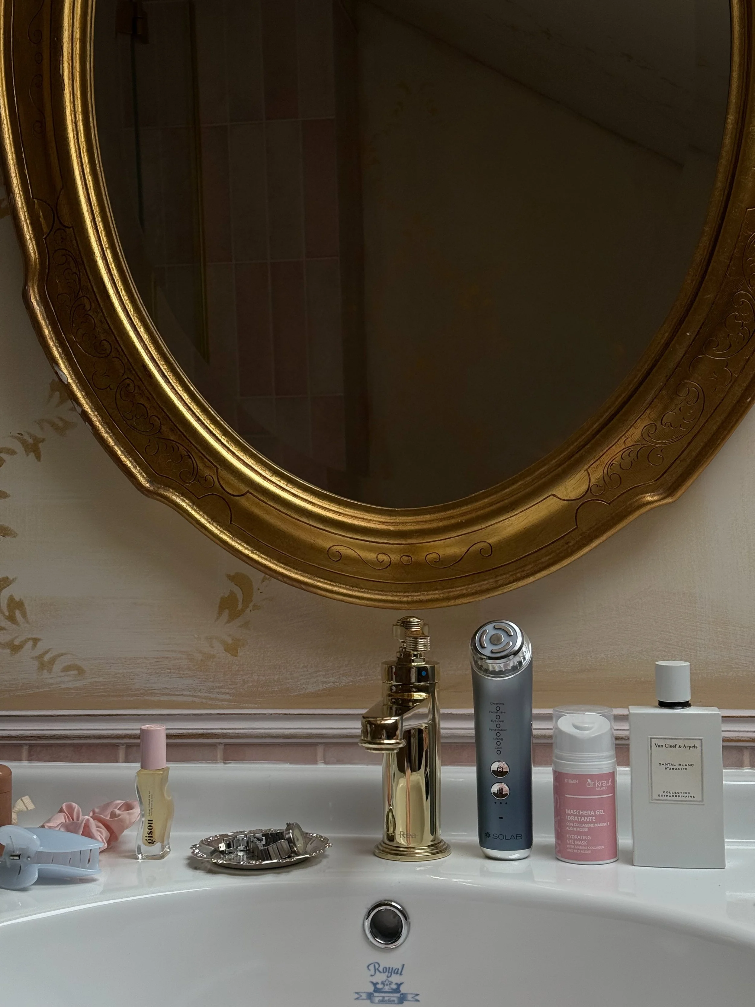 Bathroom sink with various bottles of skincare and beauty products, a silver tray with jewelry, and a large ornate gold-framed mirror above the sink.