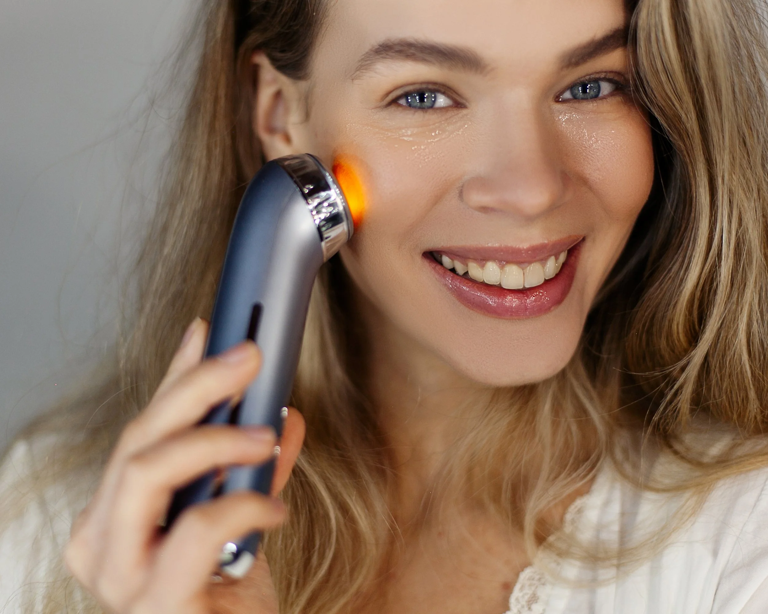 Woman with long blonde hair smiling as she uses a handheld facial skin scanner on her cheek.