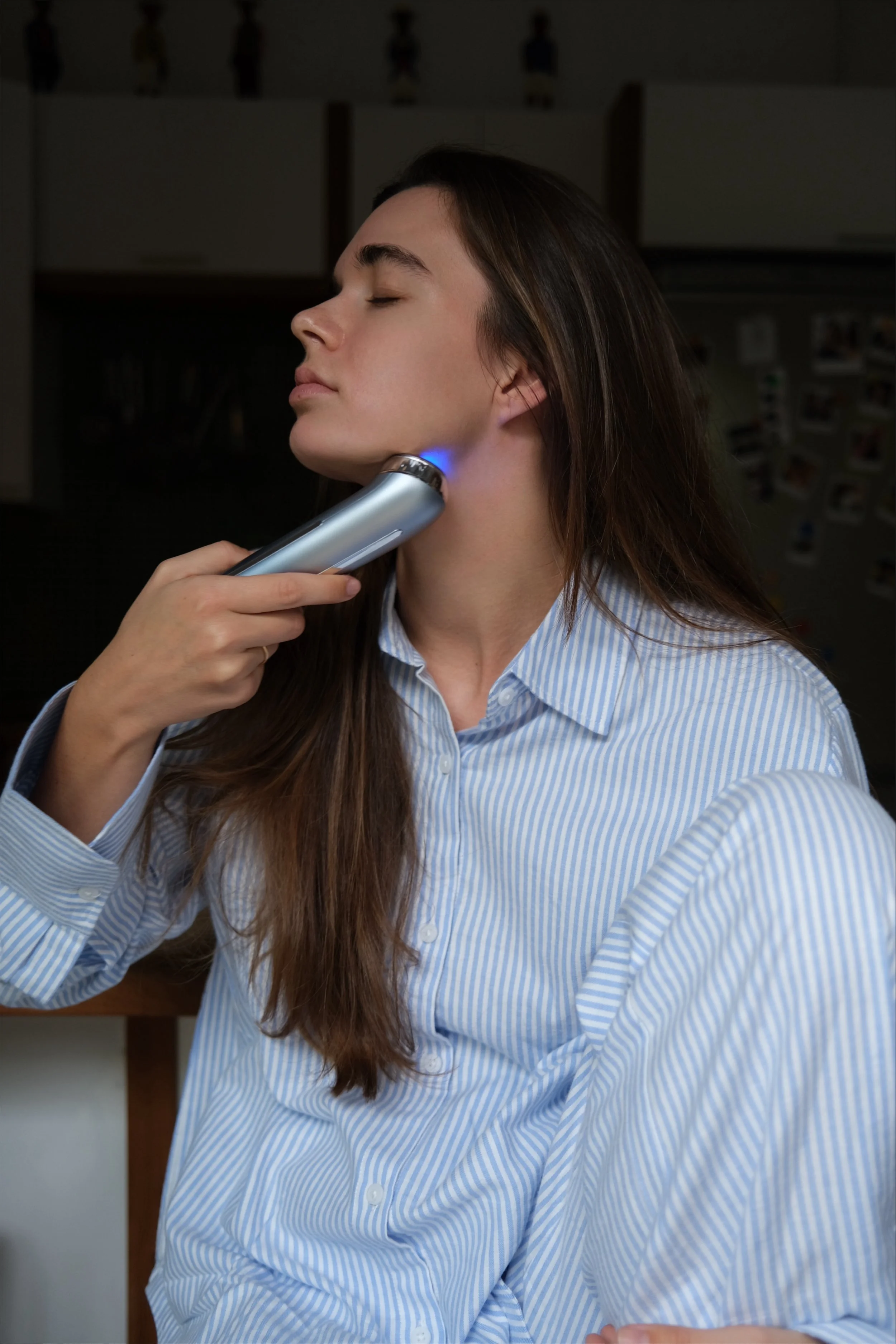 A woman with long brown hair wearing a blue and white striped shirt sits with eyes closed as a device emits a blue light to her jawline.