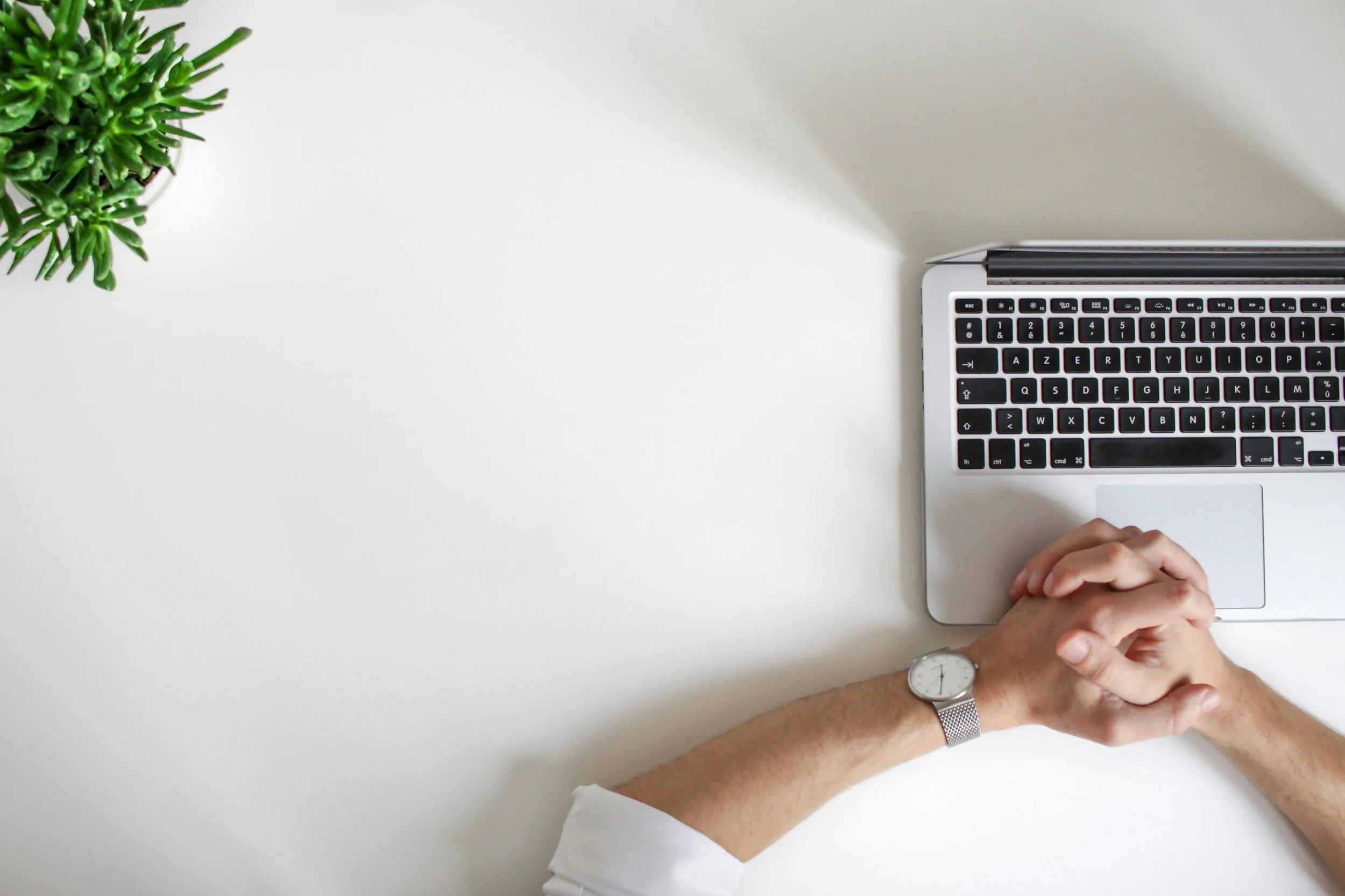 Overhead view of a person with clasped hands resting on a laptop trackpad at a white desk, with a green potted plant in the upper left corner.