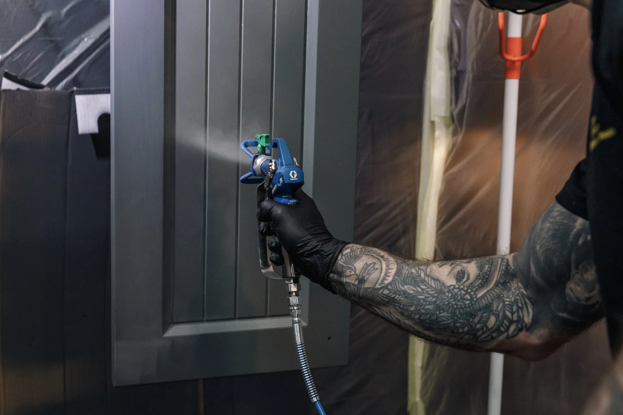 Person wearing black gloves and with tattooed arm using a spray gun to paint a metallic surface.