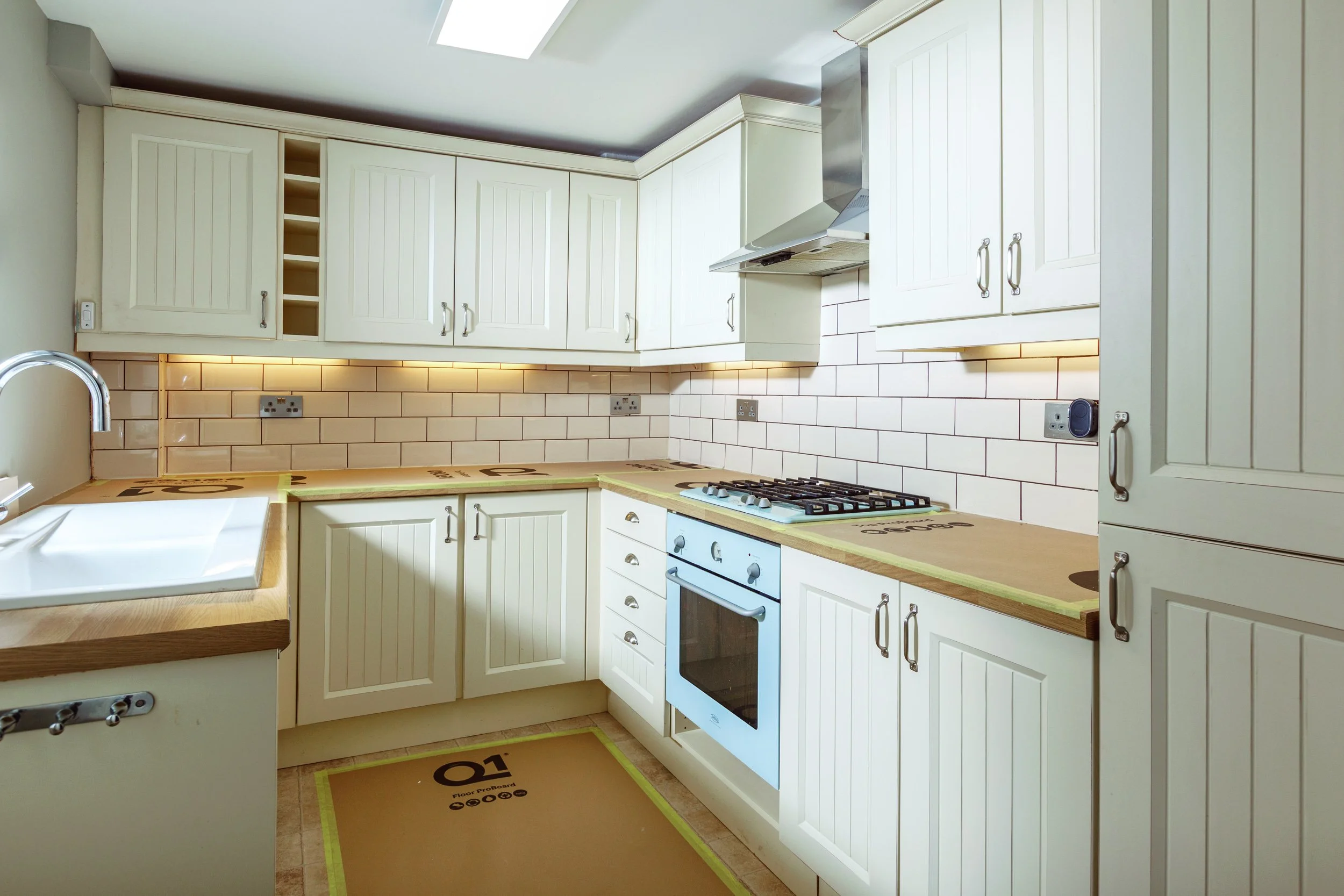 Empty kitchen with white cabinets, beige tiled backsplash, wooden countertop, white sink, stovetop, oven, and built-in storage.