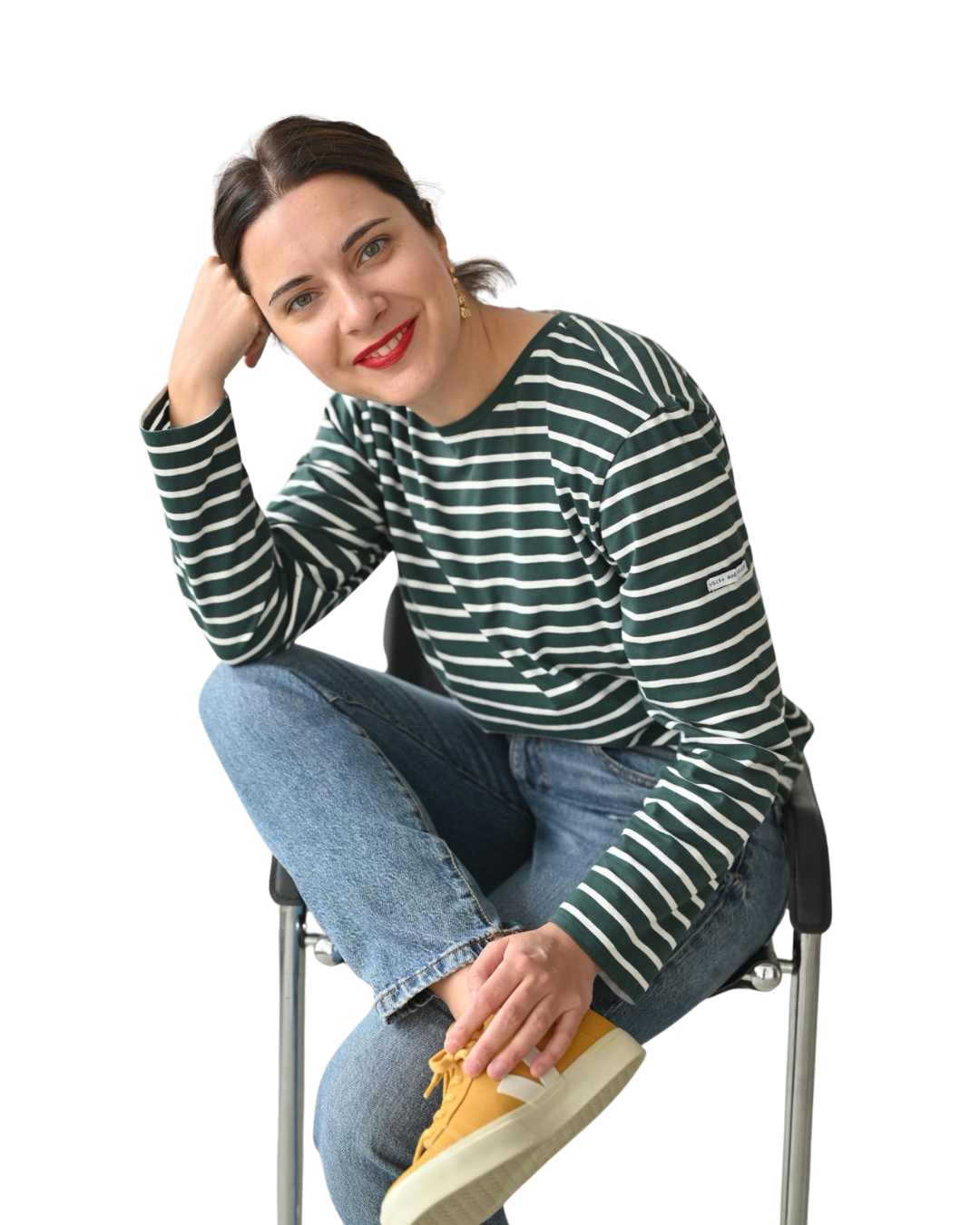 A young woman with dark hair and red lipstick, wearing a black and white striped long-sleeve shirt and blue jeans, sitting on a chair, smiling, with her right elbow resting on her knee and her hand supporting her head, while her left hand is touching a yellow sneaker on her foot.